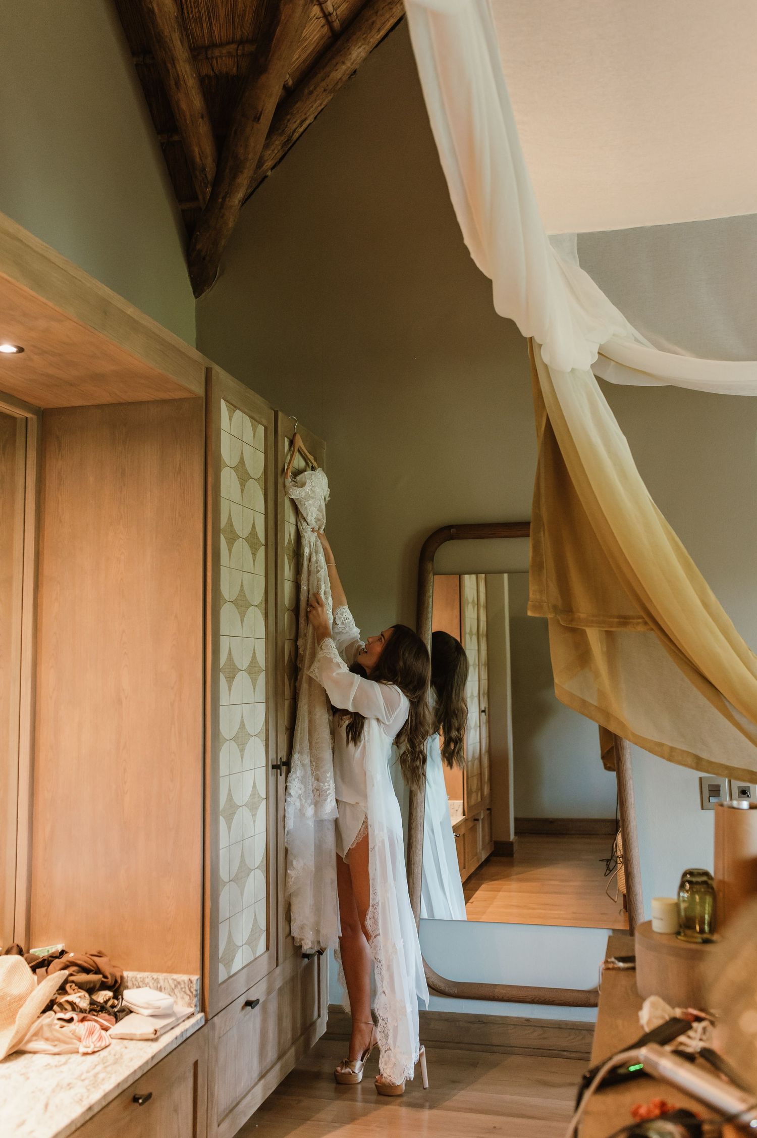 Bride in white lace robe reaching for wedding dress in elegant safari lodge dressing room.