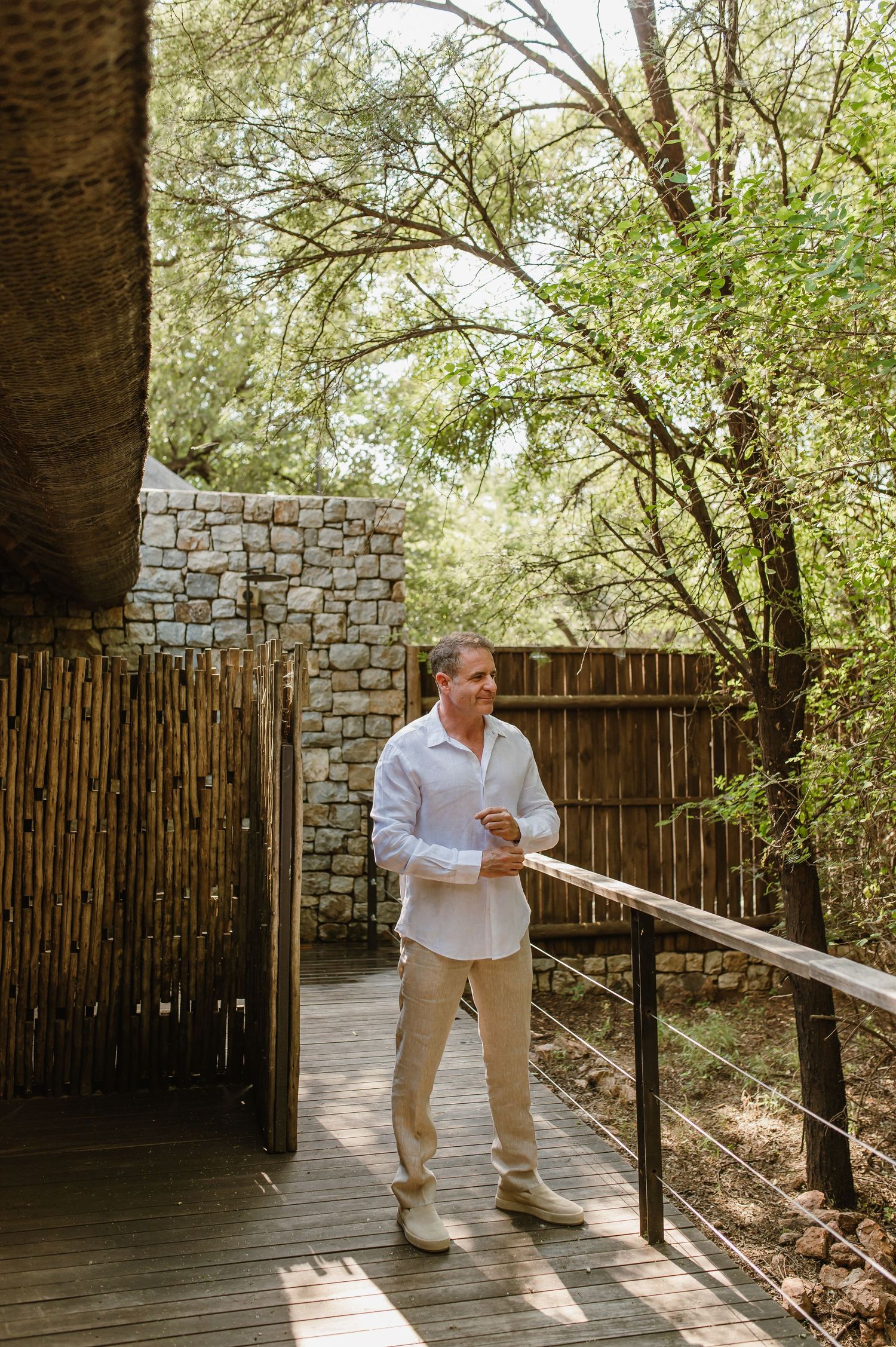 Man in white linen shirt and beige pants standing on wooden deck surrounded by bamboo fence and stone wall.