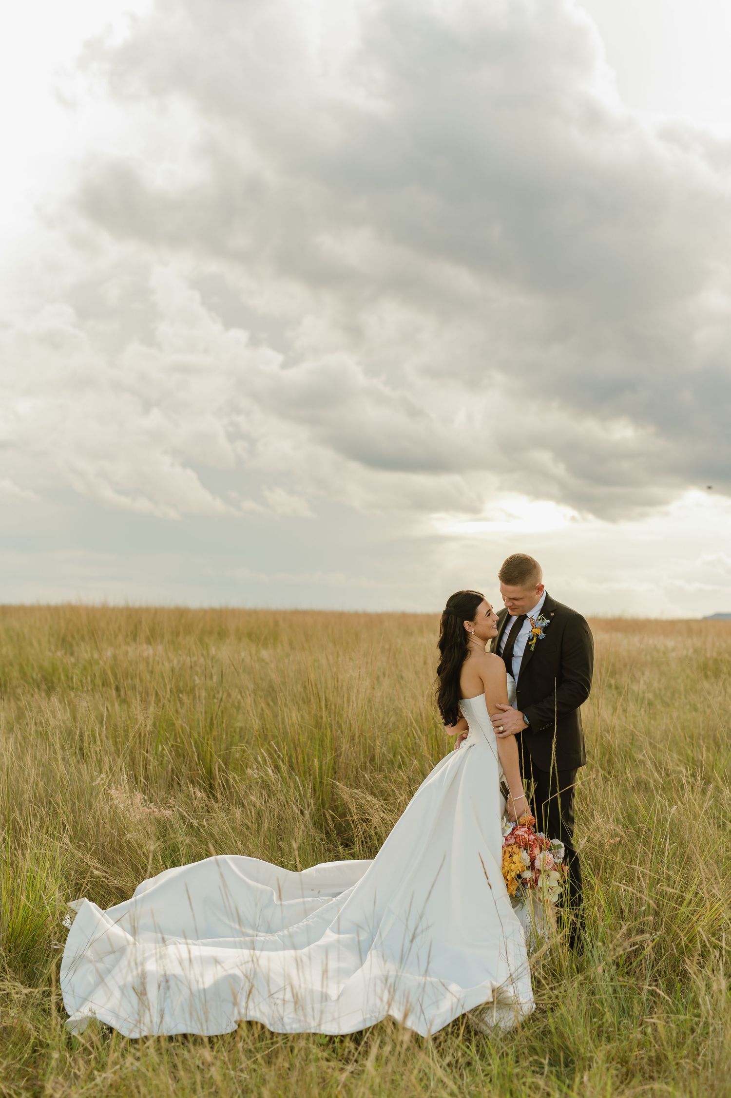 Bride and groom embrace romantically in golden meadow, her white gown trailing, under moody storm clouds.