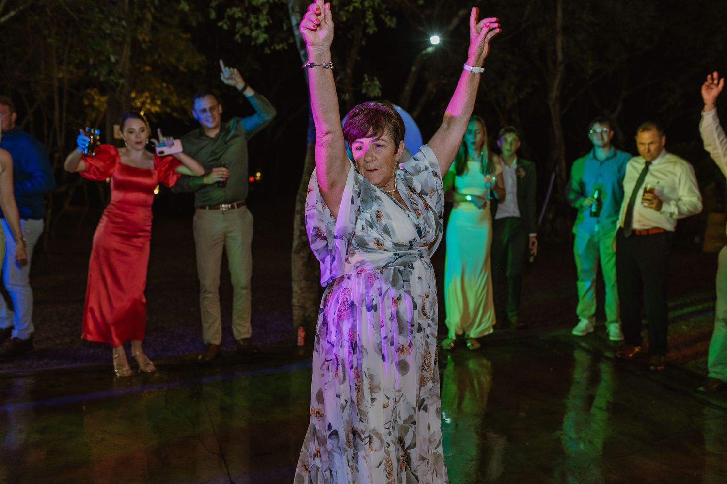Guest in floral dress dances with arms raised at a lively outdoor wedding reception at night.