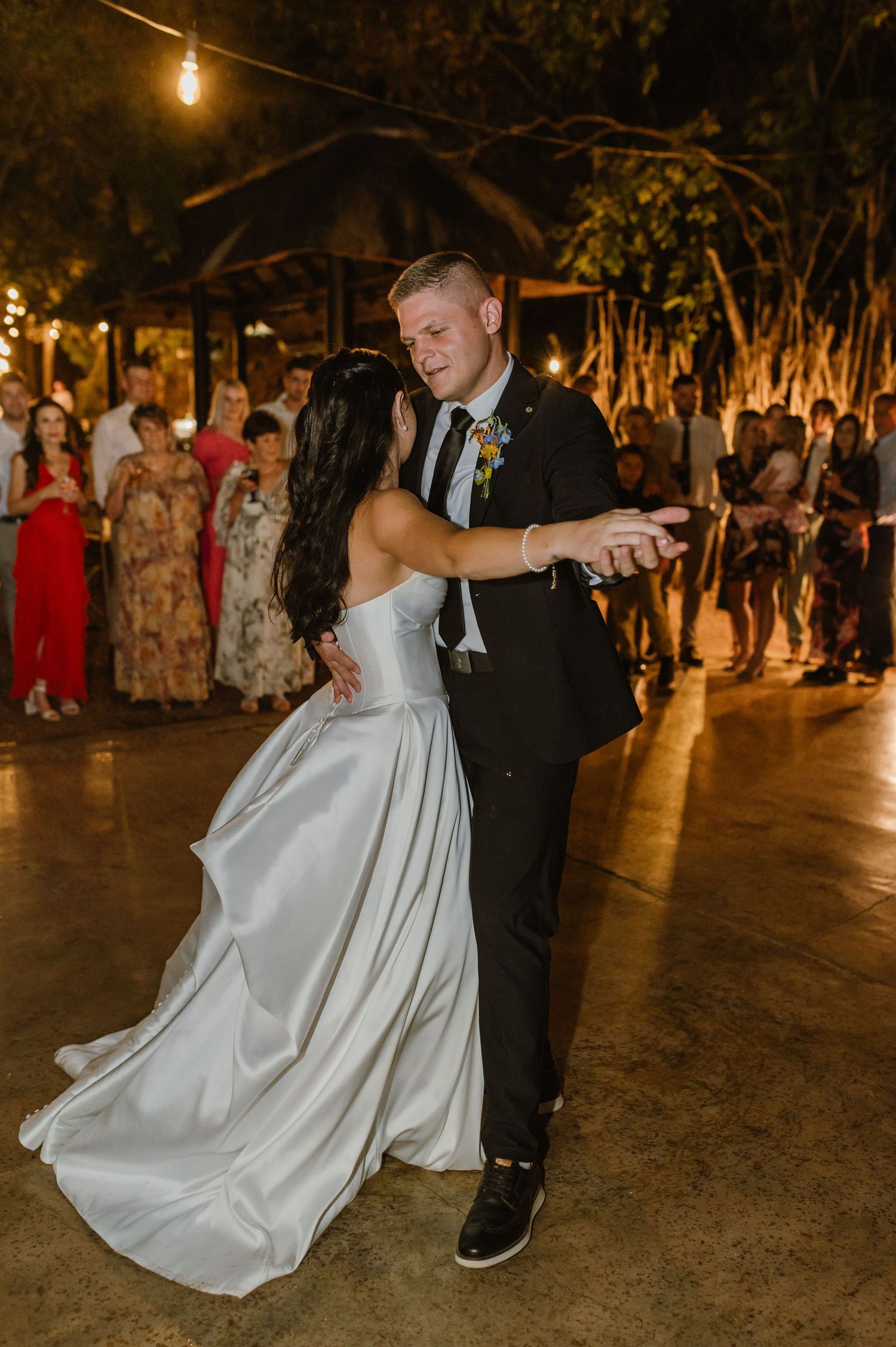 Bride and groom share their first dance at outdoor wedding reception surrounded by guests under warm string lights.