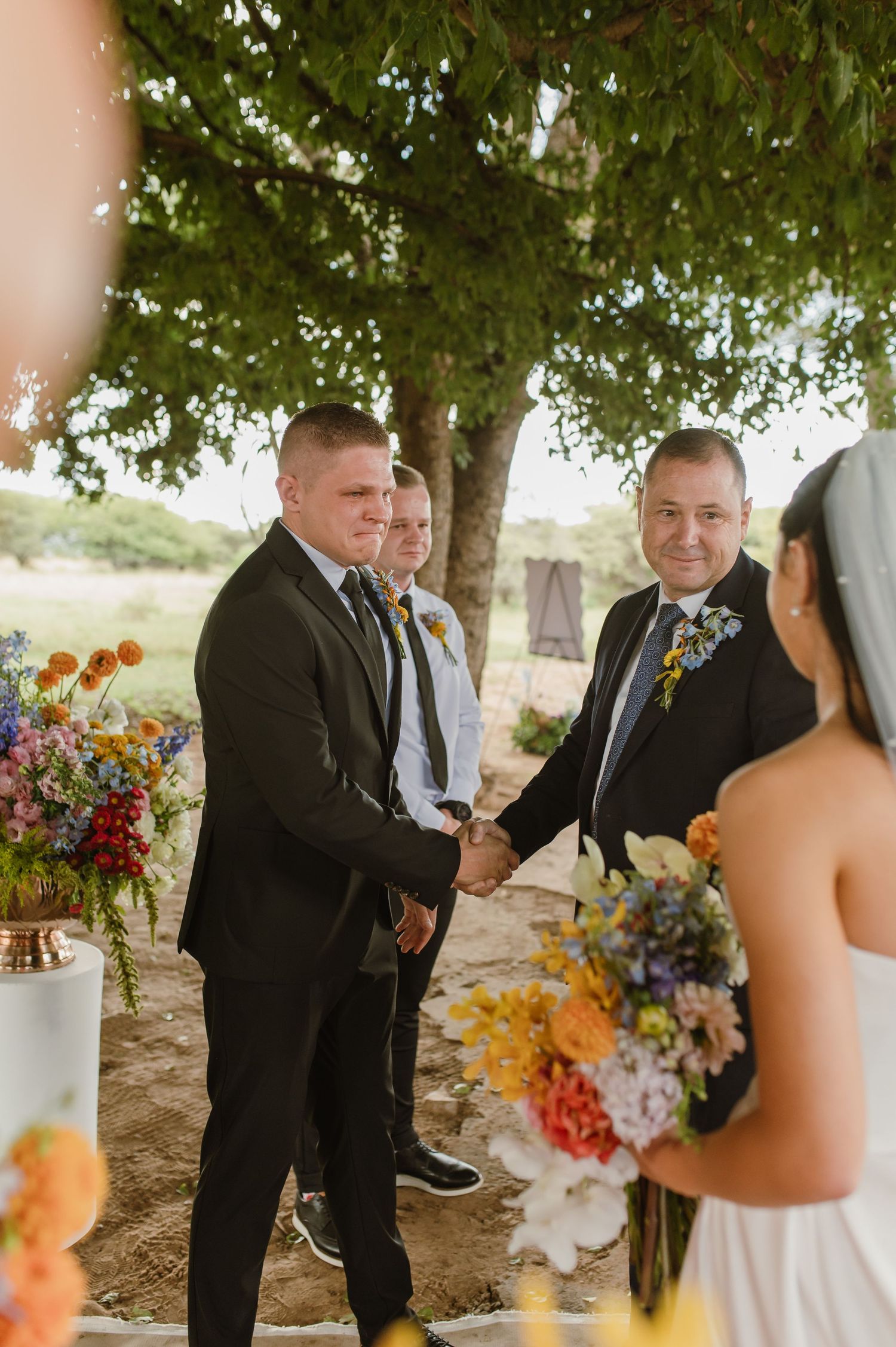 Groom shaking hands with father of the bride at outdoor wedding ceremony under a tree with colorful floral arrangements.