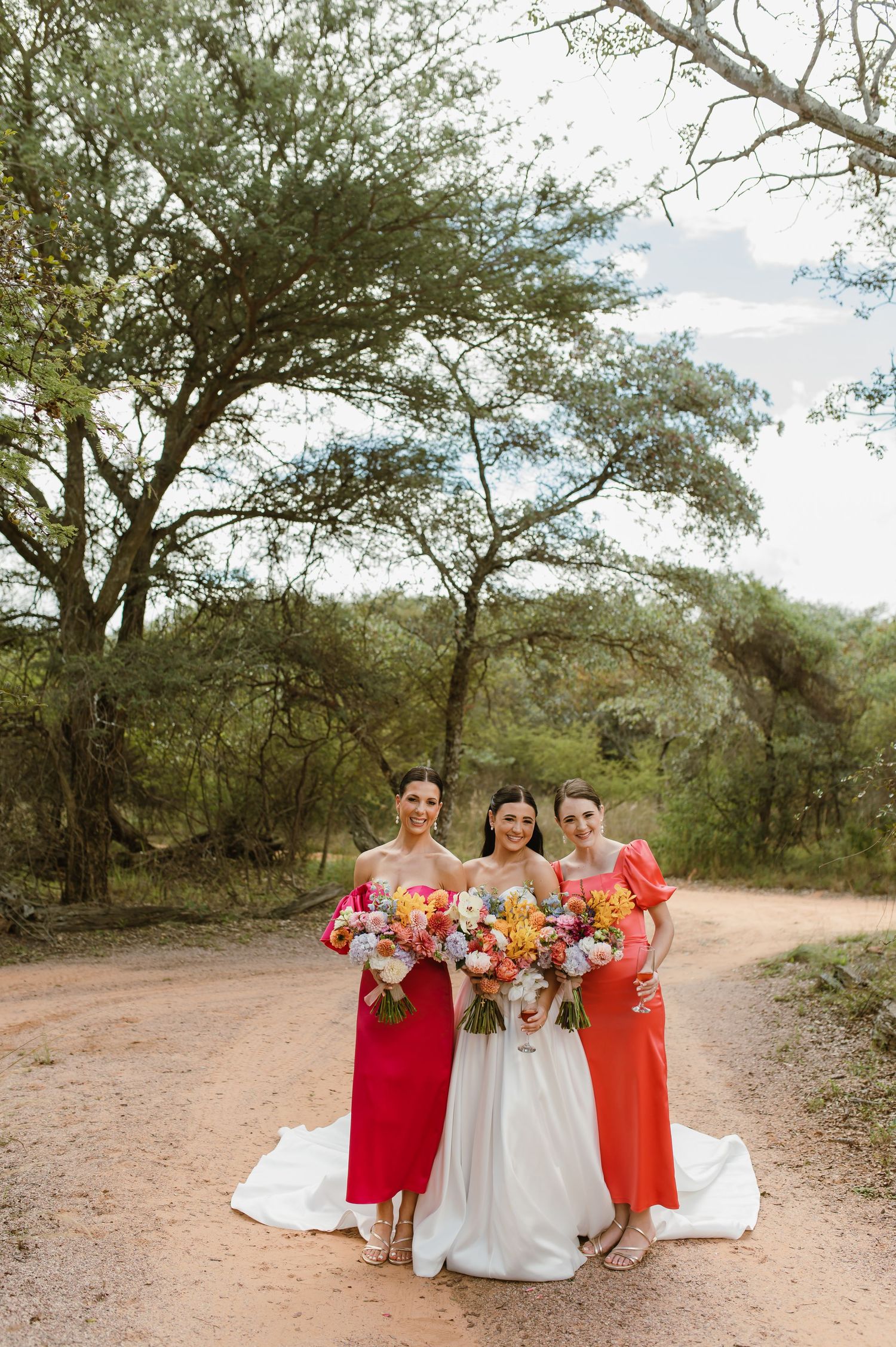 Bride in white gown posing with two bridesmaids in red and orange dresses on a bush dirt road.