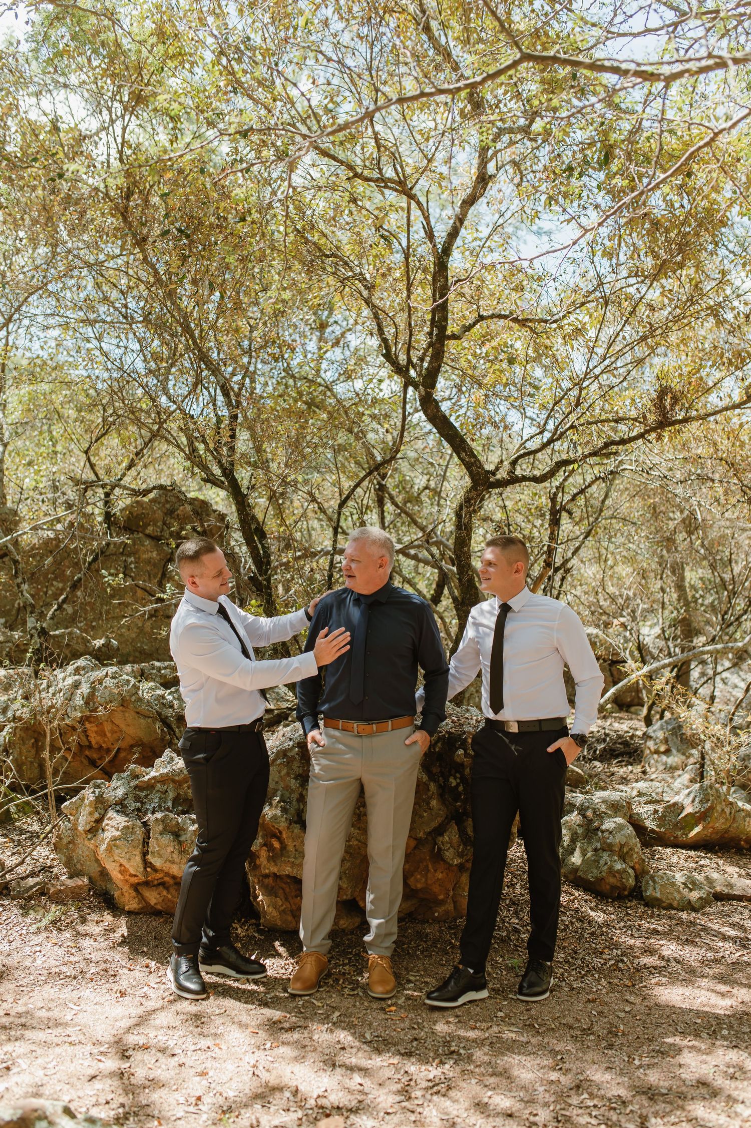 Three men in formal attire share a moment outdoors among rocky terrain and trees.