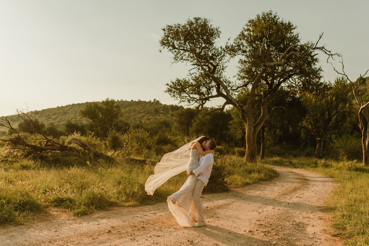 A couple shares a romantic kiss on a dirt path surrounded by lush greenery, with a flowing veil caught in the breeze.