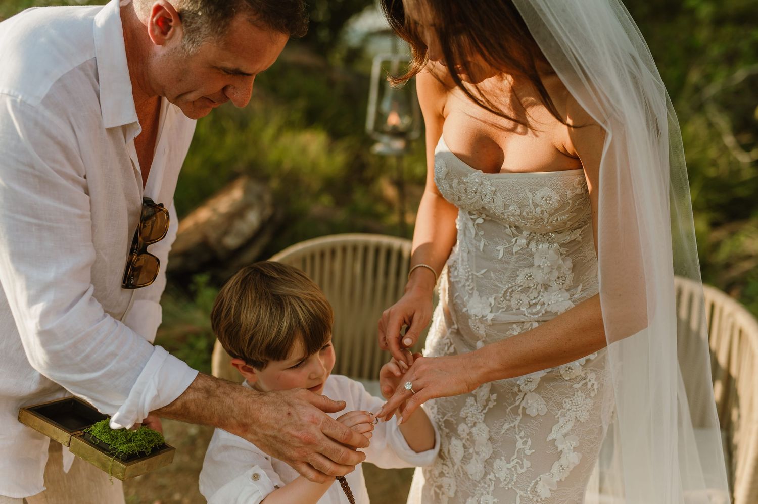 A groom and bride in a lace wedding dress attend to a young child during an outdoor wedding ceremony.