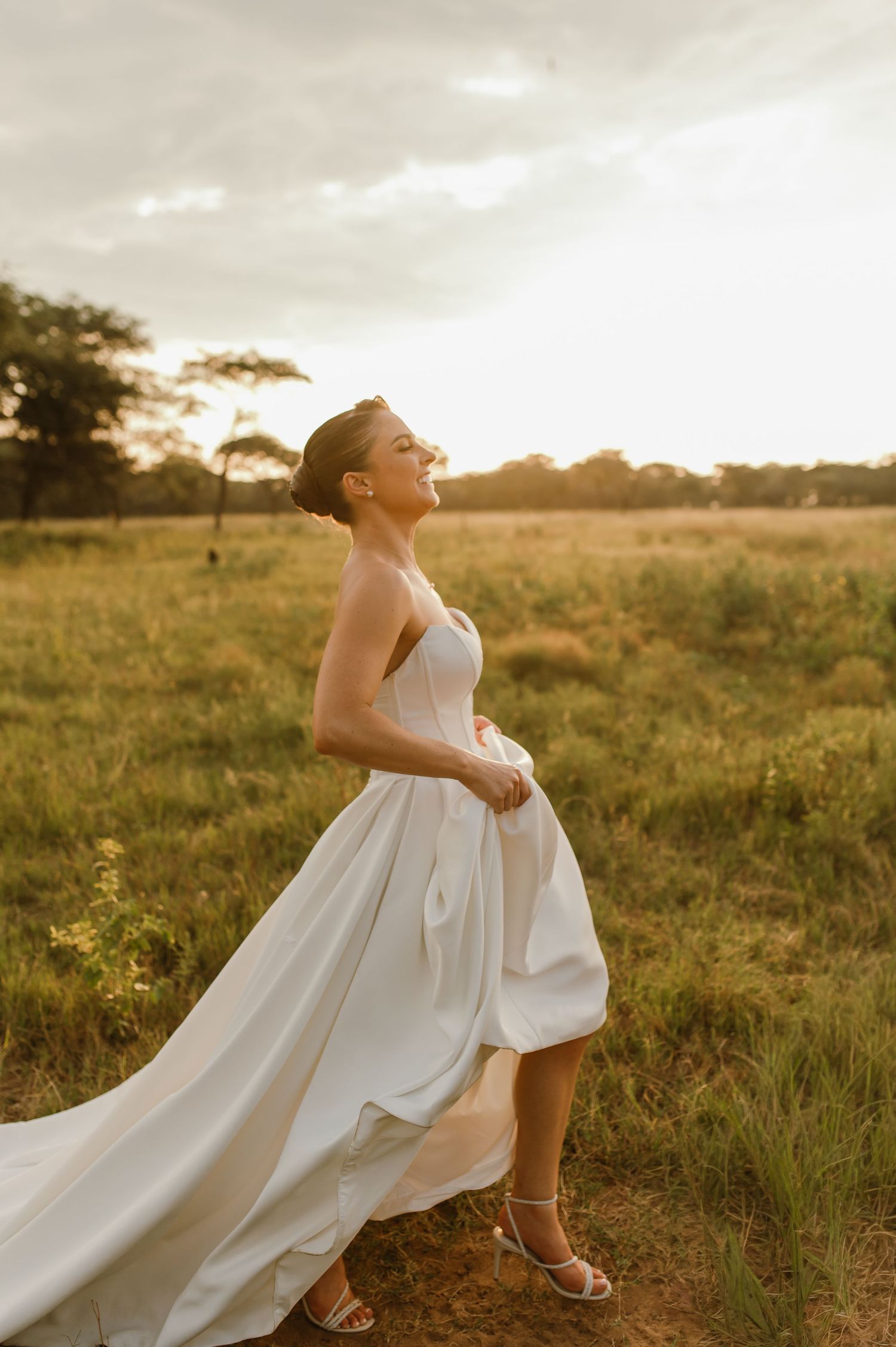A flowing white wedding dress billows in the breeze as someone stands in a golden field at sunset.