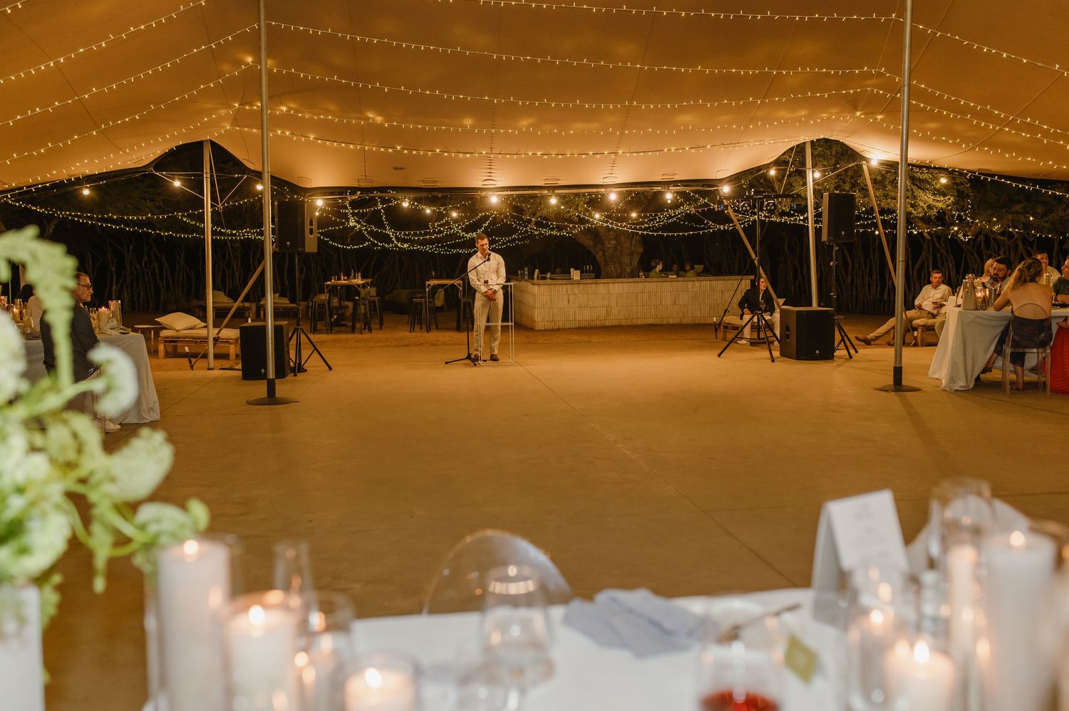 Speaker addressing guests at an elegant tented evening reception with candlelit tables.