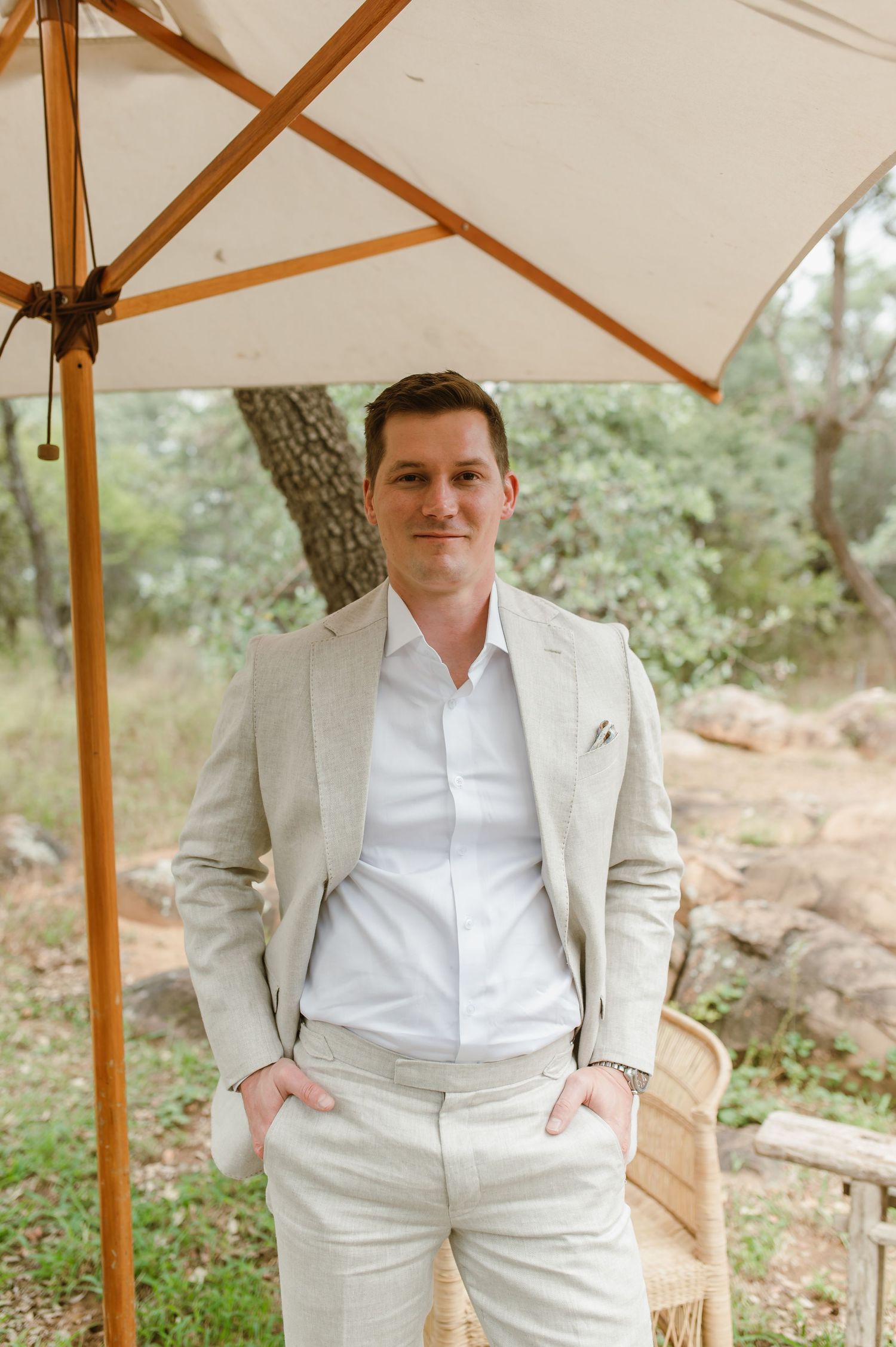 A person in a light beige suit and white shirt stands under a cream-colored umbrella in an outdoor natural setting.