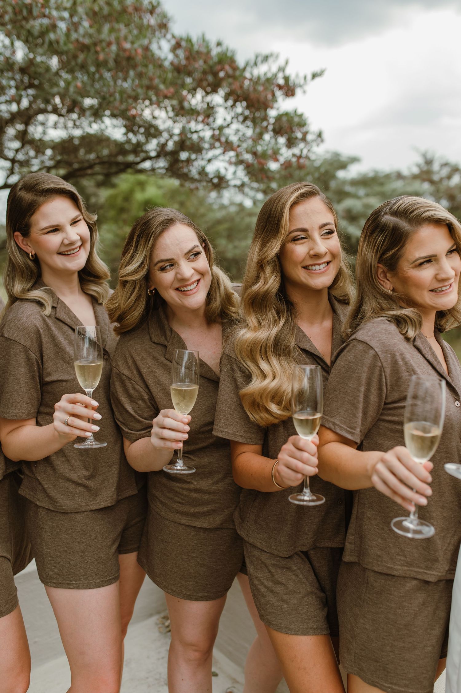 Four women in matching brown outfits holding champagne glasses and smiling outdoors.