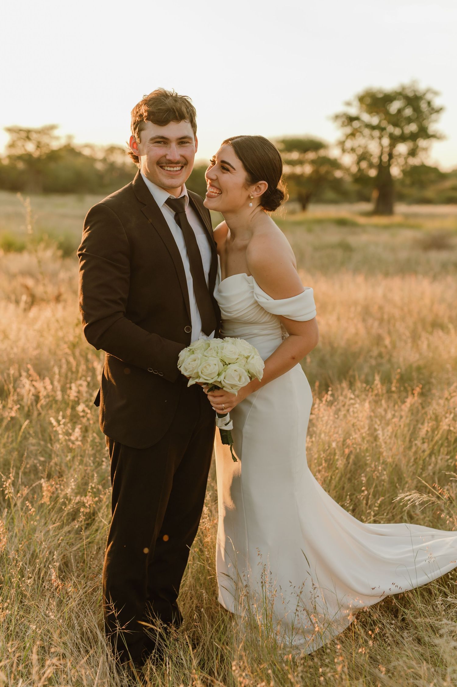 A smiling bride and groom pose in a golden sunlit field, she holds white roses in an off-shoulder gown, he wears a black suit.
