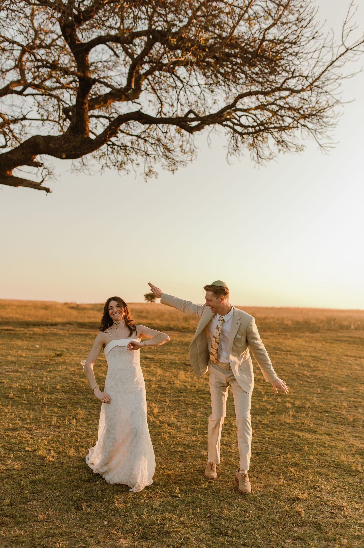 Bride and groom dancing joyfully in golden field at sunset beneath a bare oak tree.