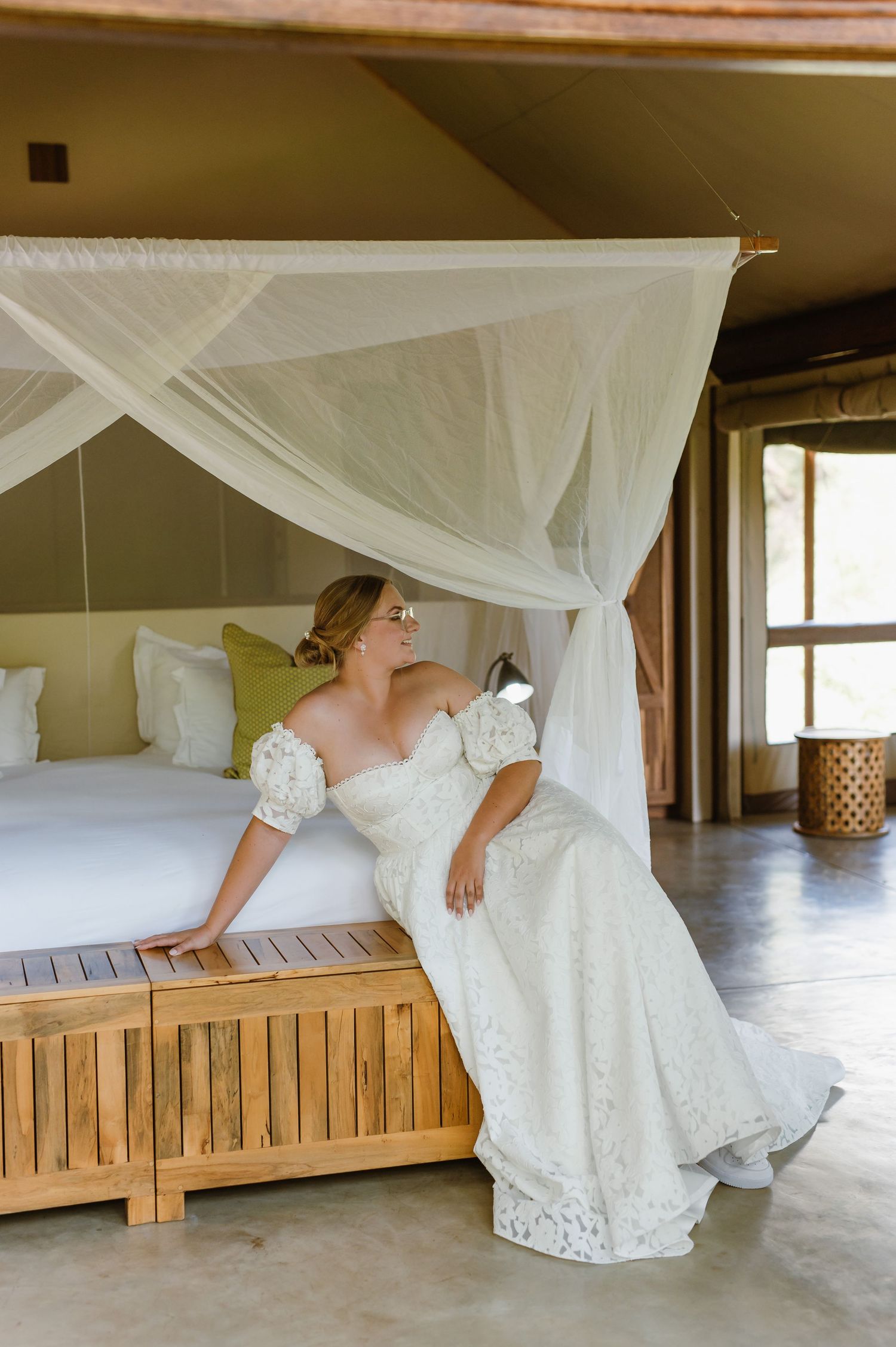 A white wedding dress flows elegantly across a wooden bench under sheer canopy draping in a sunlit room.