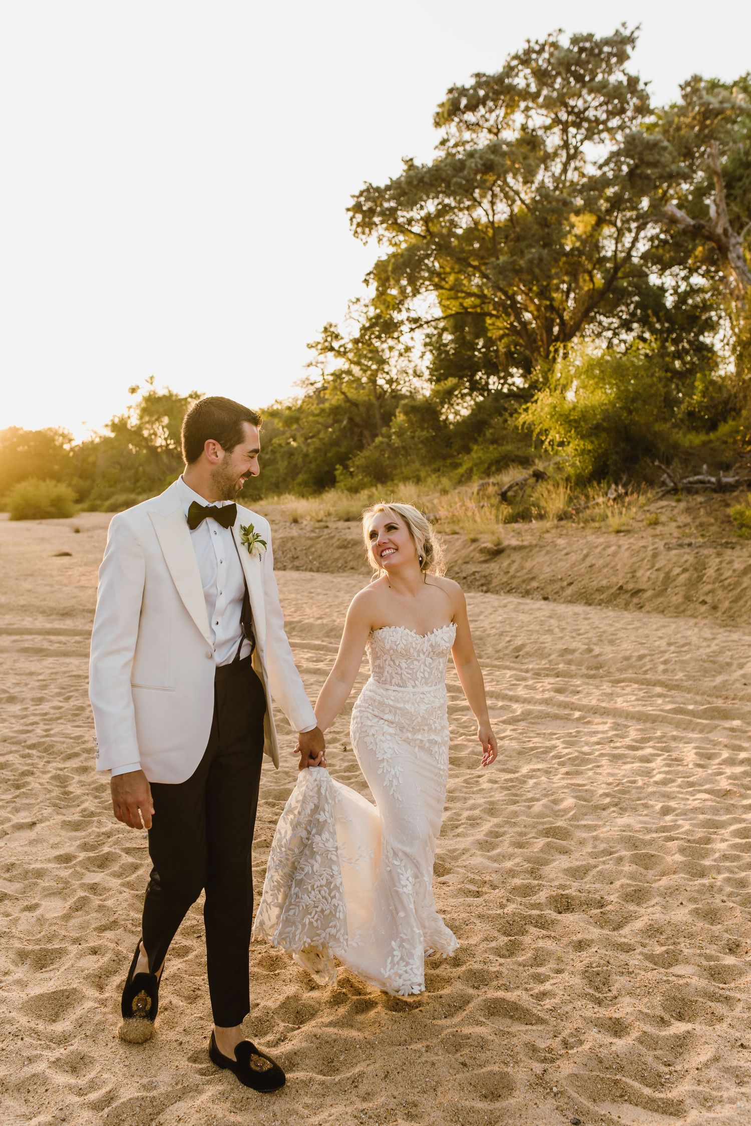 Couple in wedding attire walking together on sandy beach during golden hour sunset.