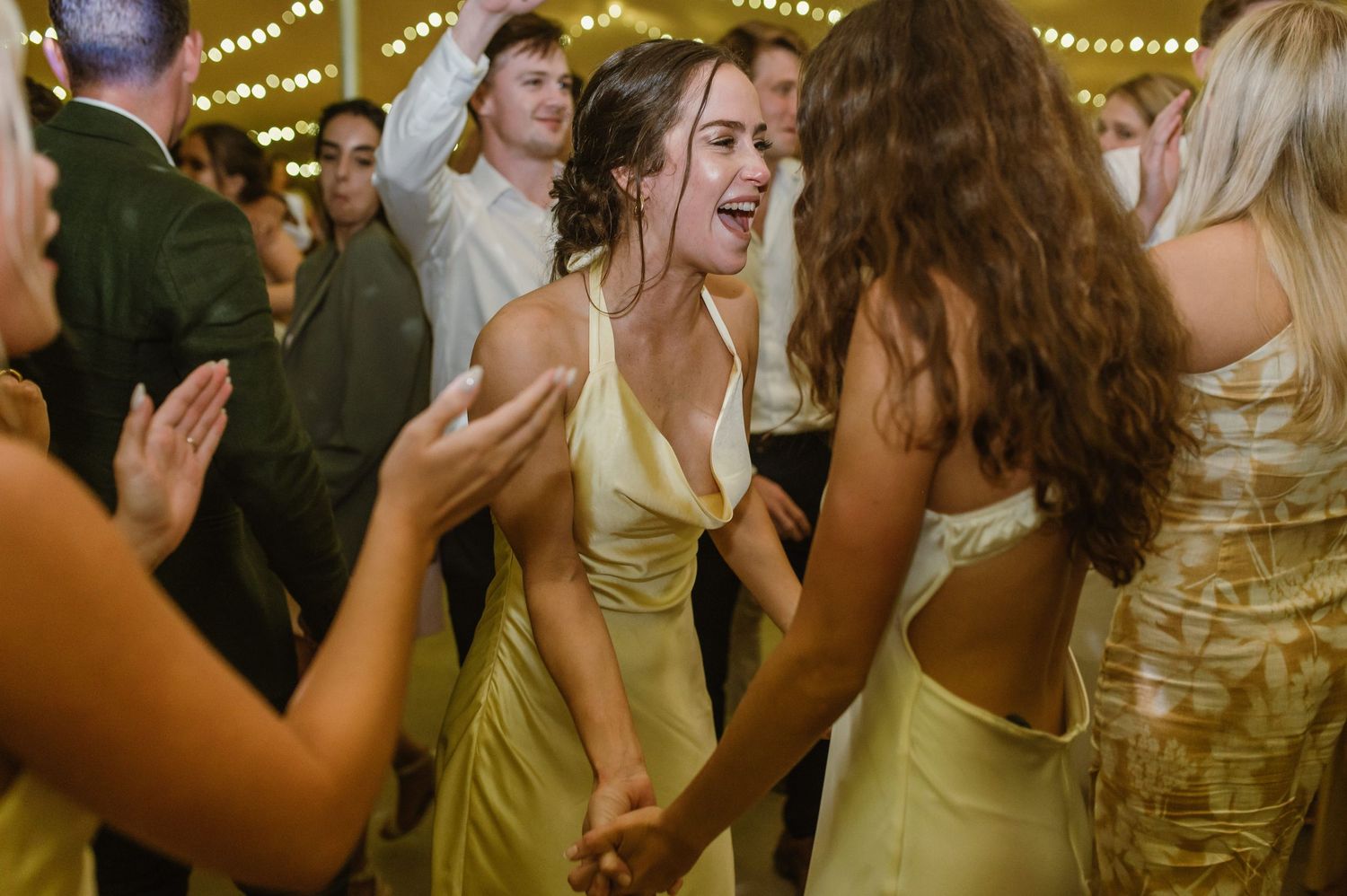 Wedding party members dancing and laughing together under decorative string lighting.