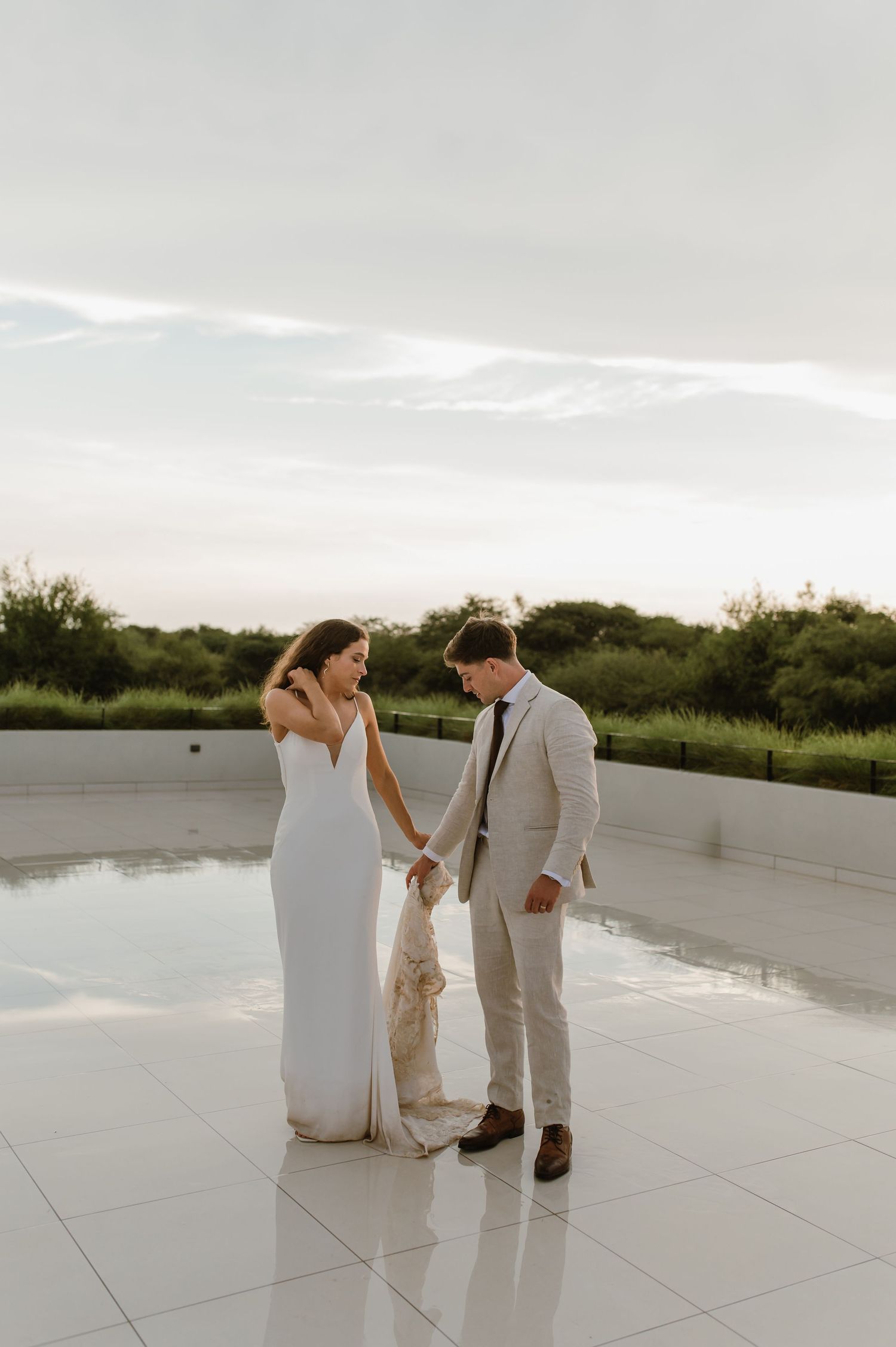 A wedding couple stands together on a reflective white surface with greenery in the background.