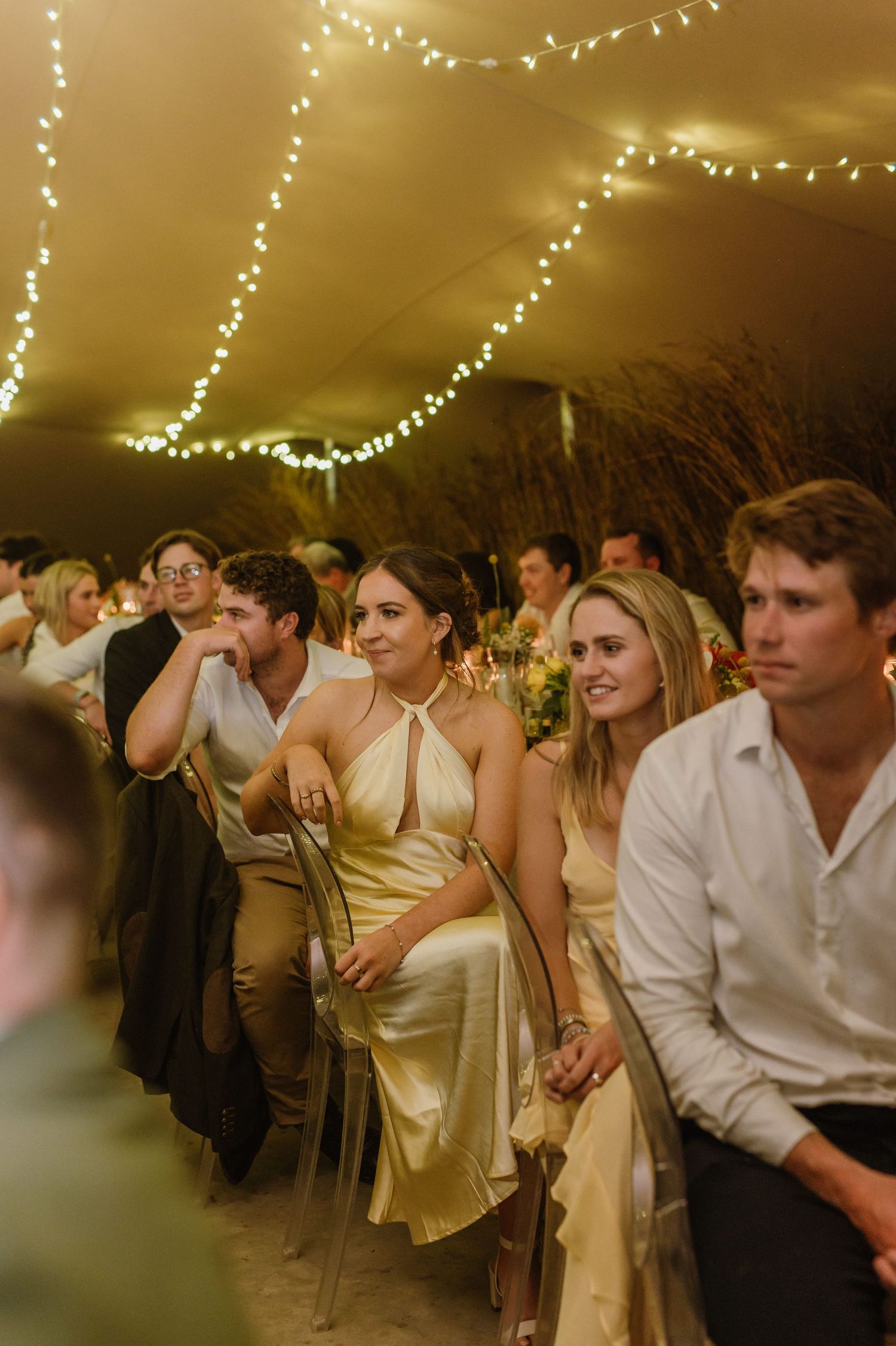 Wedding guests seated at tables in warm lighting with string lights creating an intimate atmosphere.