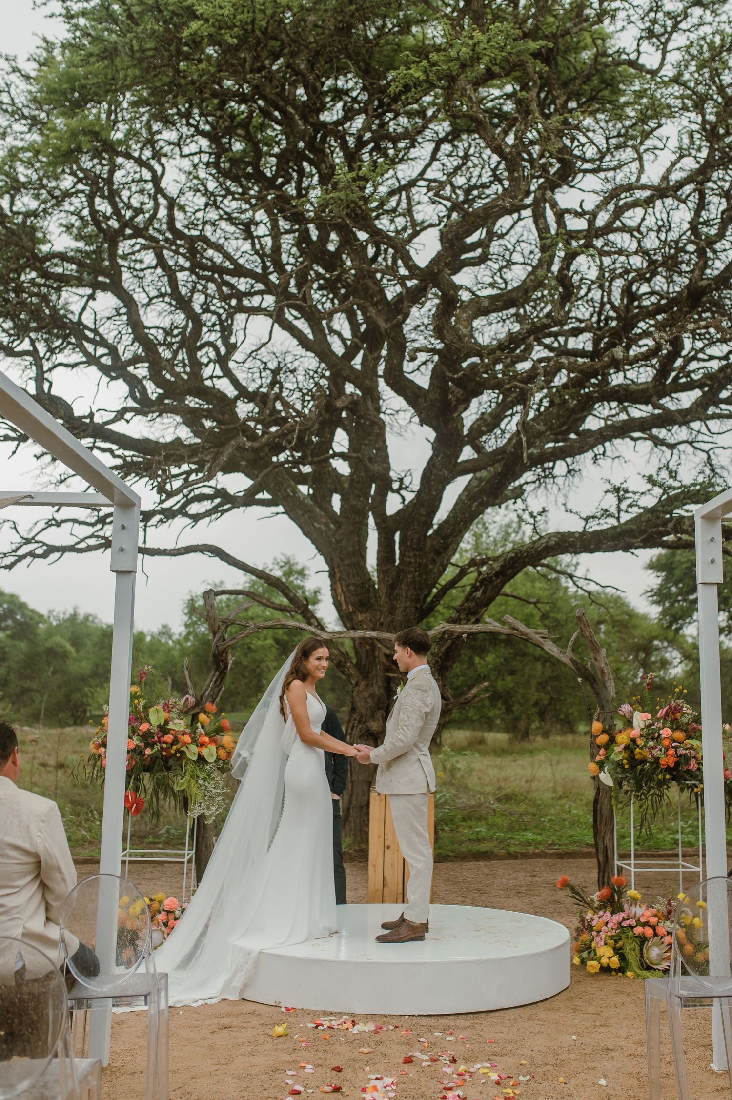 A wedding ceremony on a circular platform beneath a majestic tree with floral decorations.