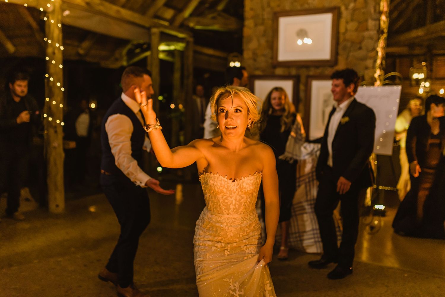 Wedding celebration in a barn venue with guests dancing under string lights and wooden rafters.