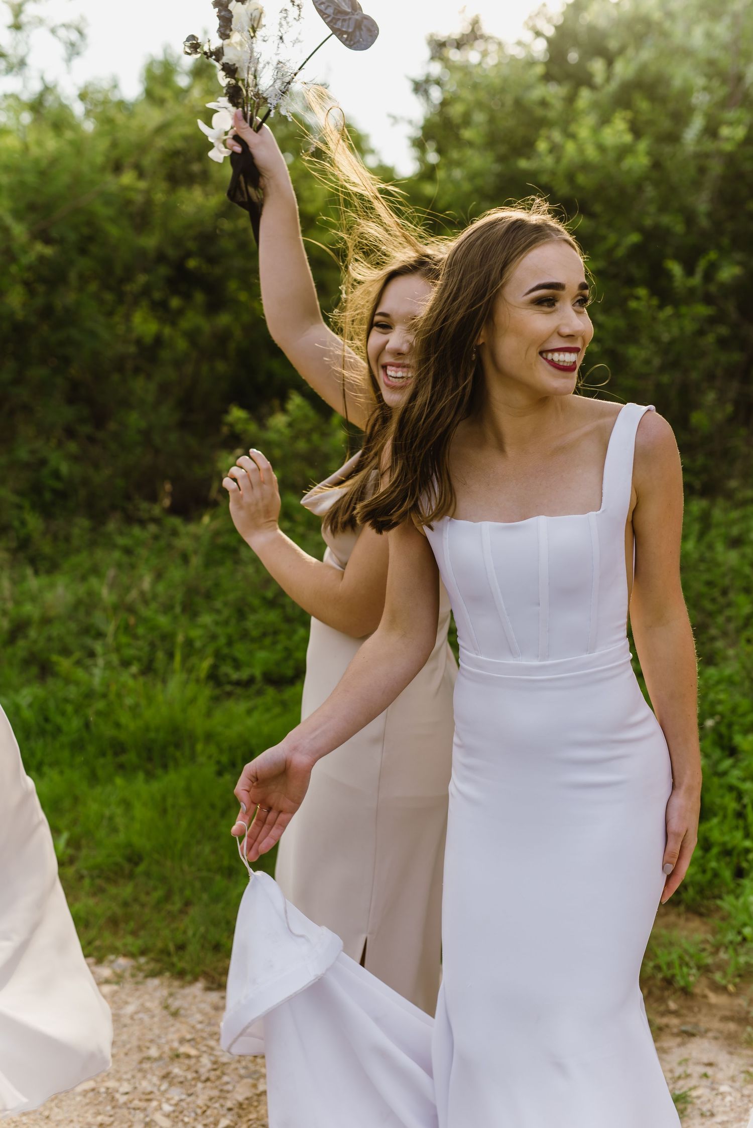 Two people in white dresses dance joyfully together on a dirt path surrounded by greenery.