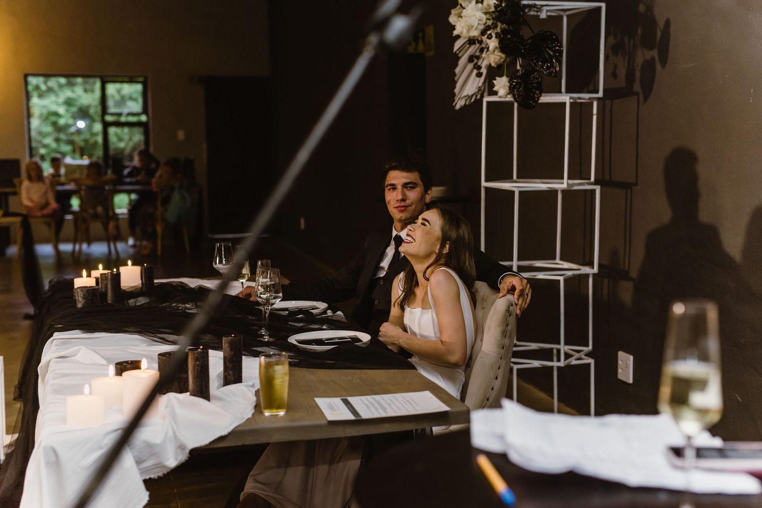 A couple sits at their reception table during speeches at an indoor venue.