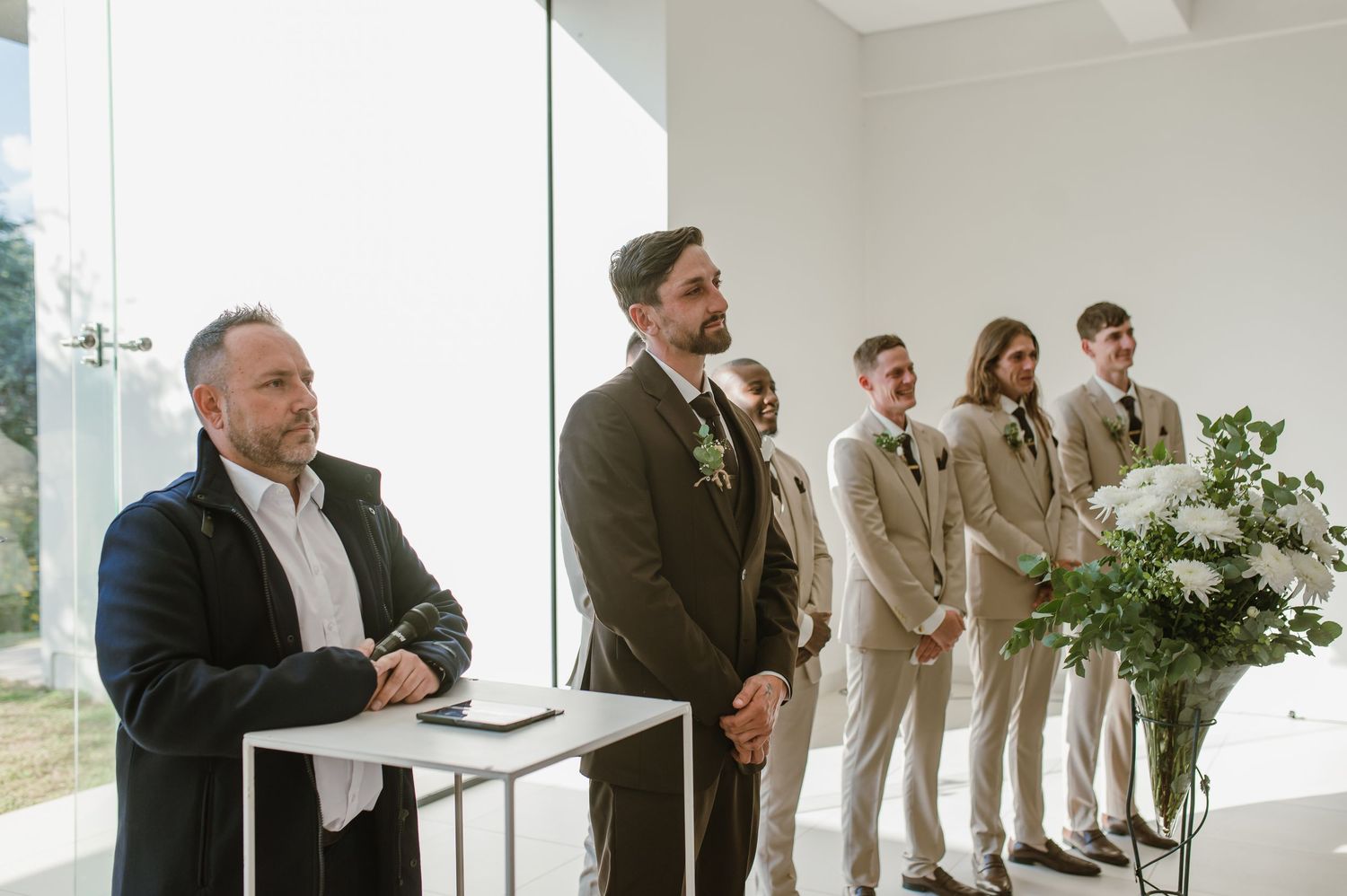 Wedding party lined up inside modern white venue with large windows and natural decor.
