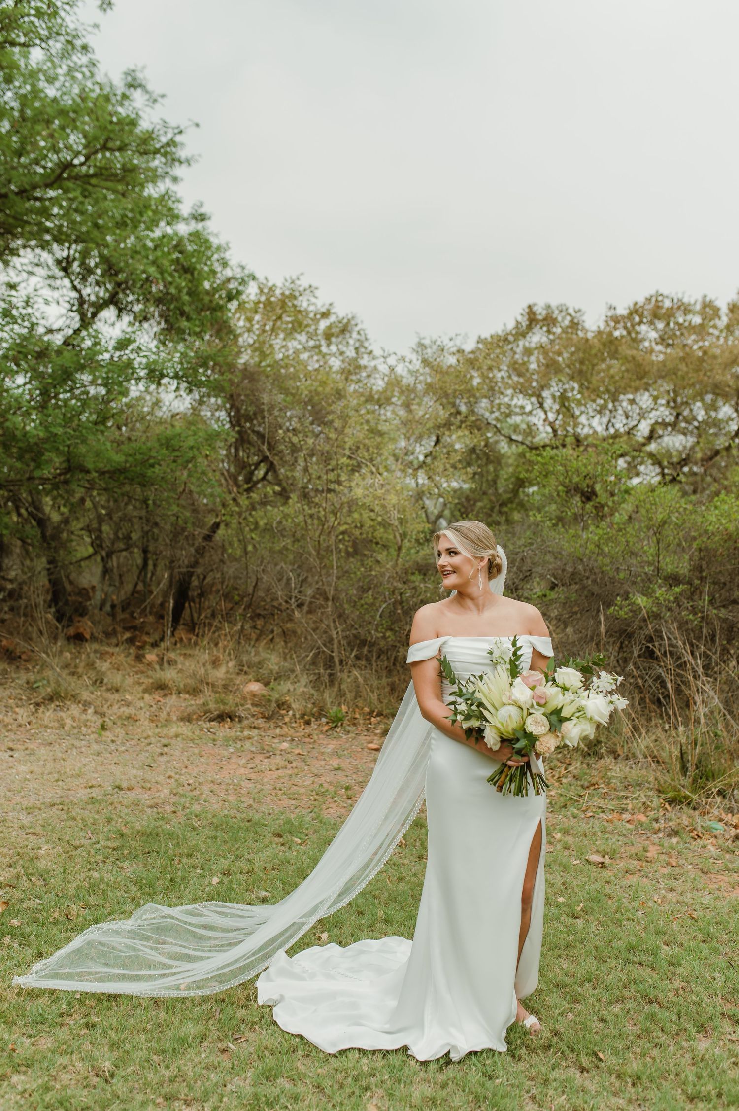 Bride holds white bouquet while her veil flows in the breeze against natural outdoor backdrop.