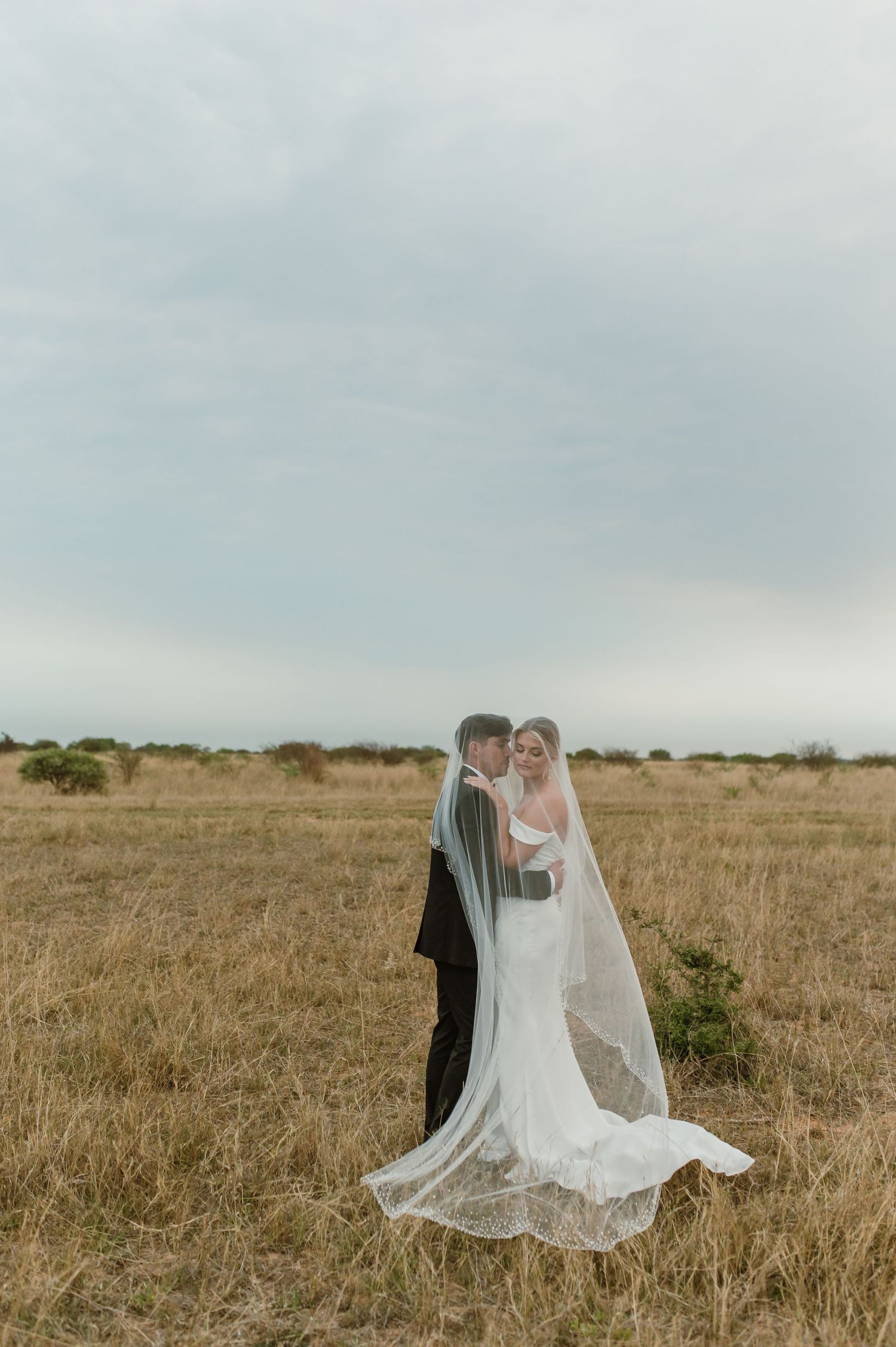 Bride and groom share romantic moment under flowing veil in countryside setting.