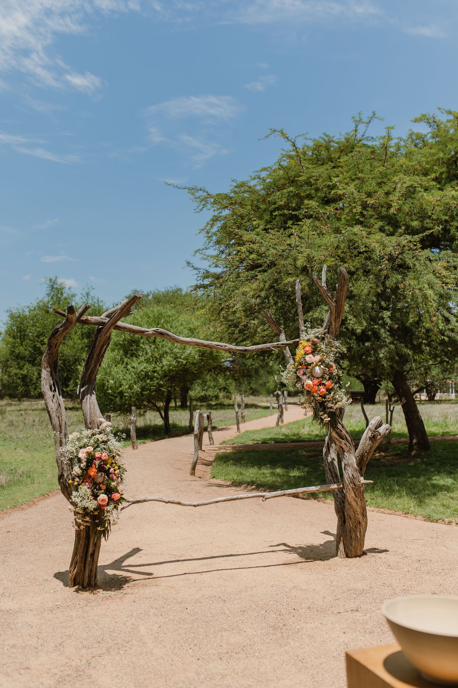 Rustic wooden wedding arch decorated with floral arrangements on a dirt path surrounded by green trees.