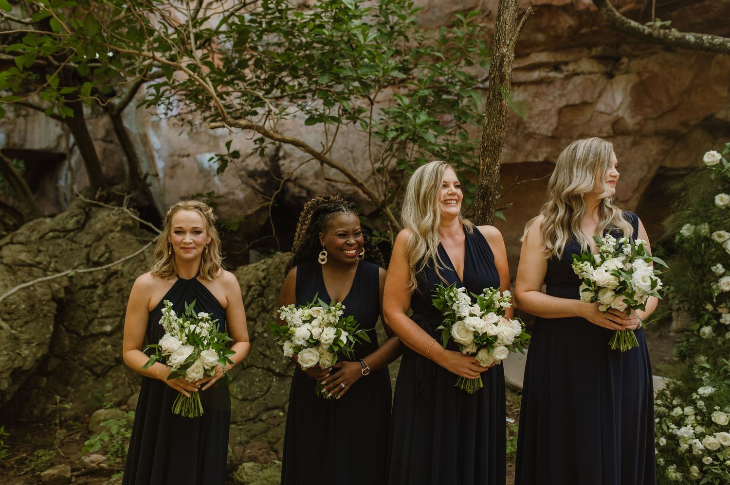Bridesmaids in navy dresses holding white floral bouquets stand in a wooded setting.
