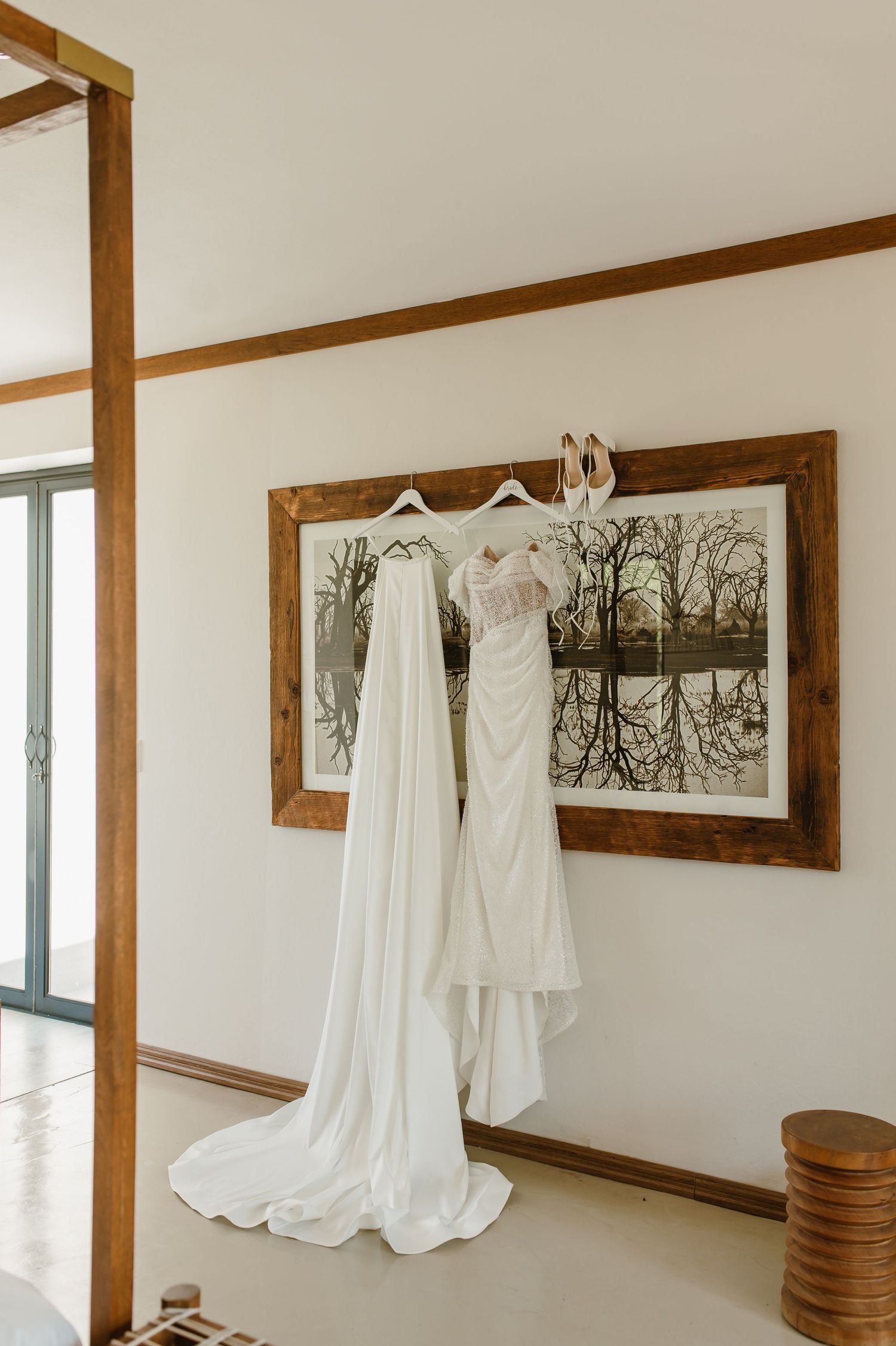 Wedding dresses displayed on hangers against white walls with rustic wooden framed artwork.