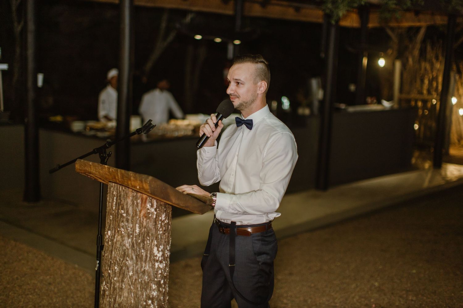 Speaker at wooden podium during outdoor evening event with string lights overhead.