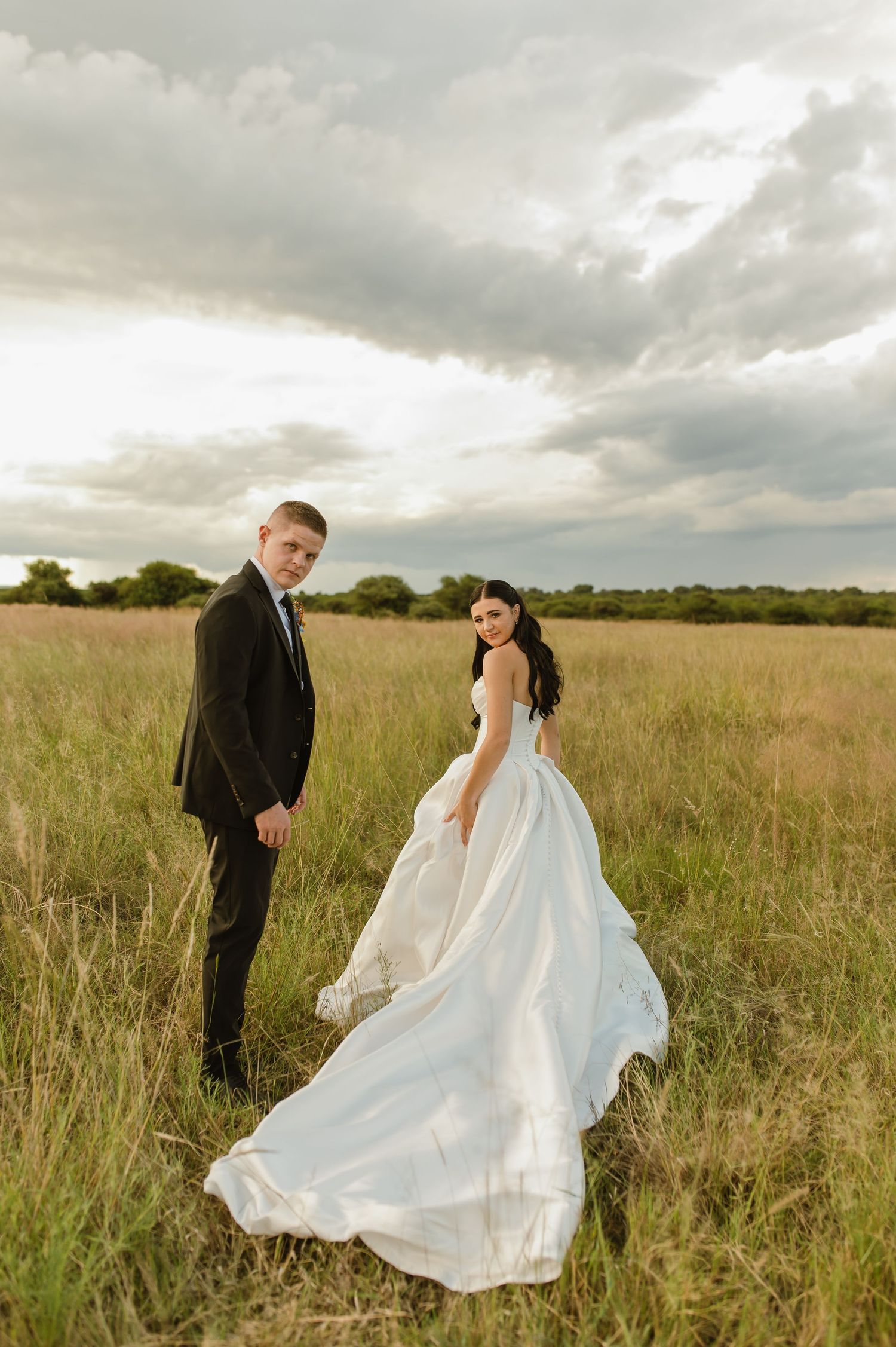 Bride in white ballgown and groom in black suit hold hands walking through sunlit grassy field.