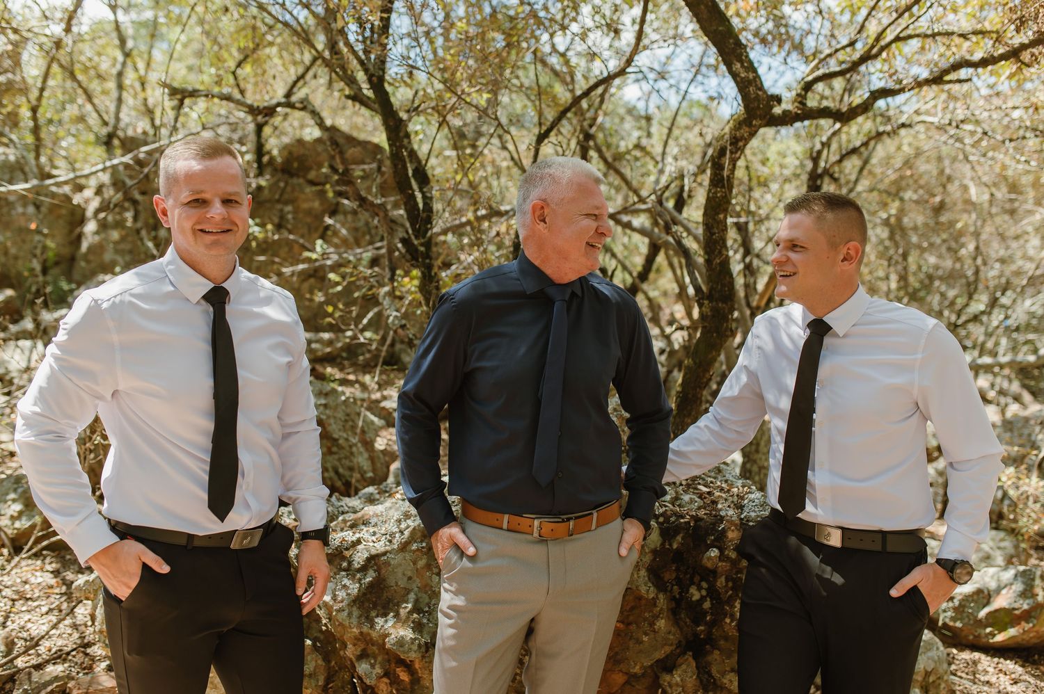 Three men in white shirts and black ties laughing outdoors among trees before wedding.