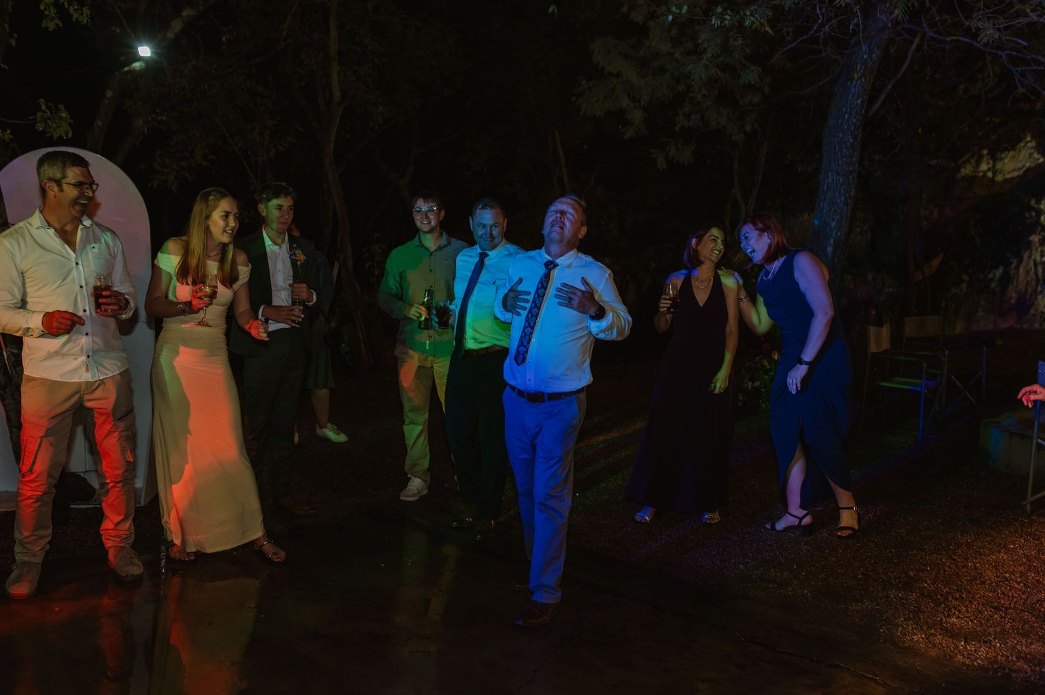 Wedding guests dance and celebrate under colorful lights at outdoor nighttime wedding reception.