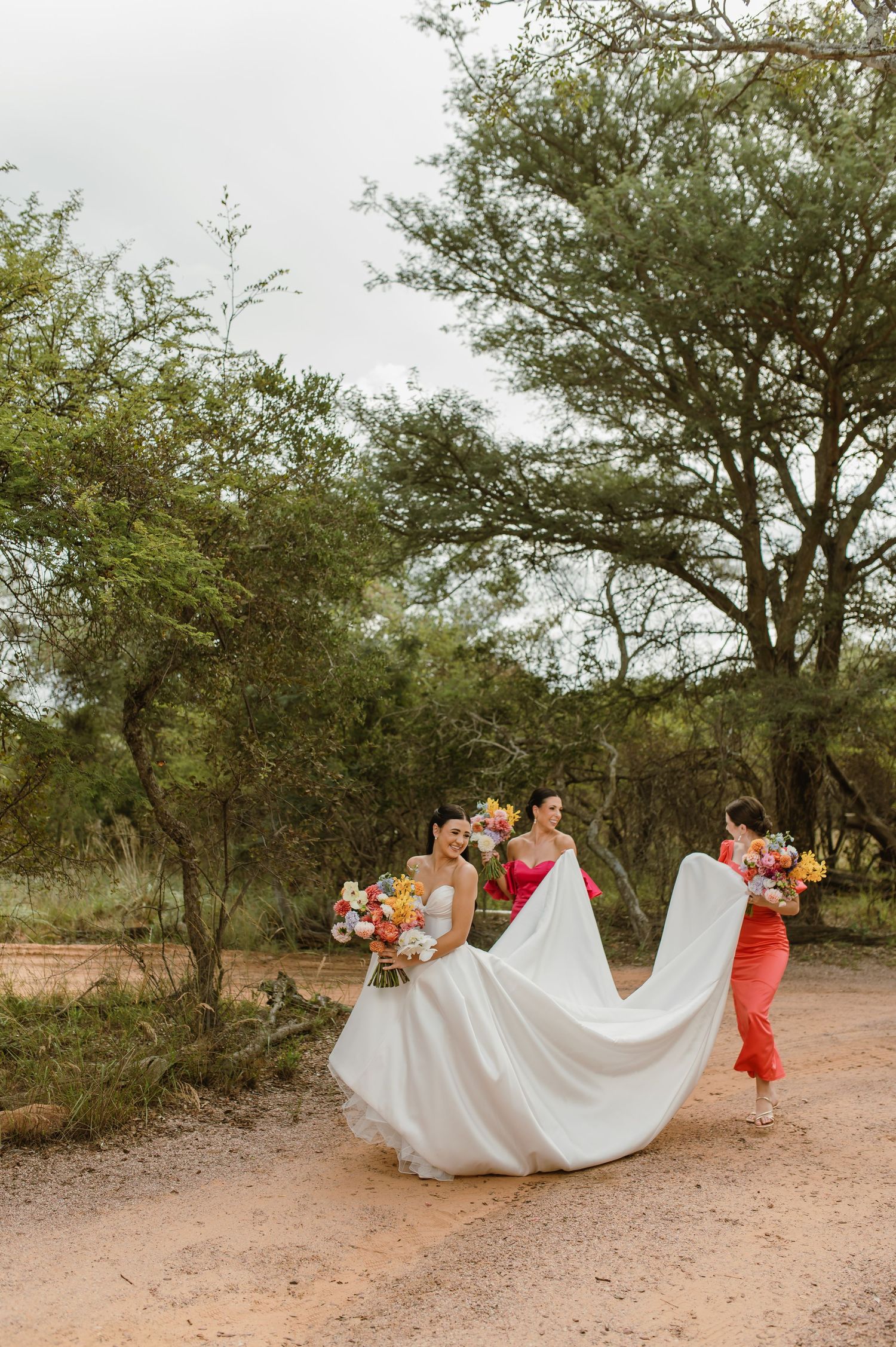 Bride walking with bridesmaids on a dirt path in the African bush, holding colorful bridal bouquets.
