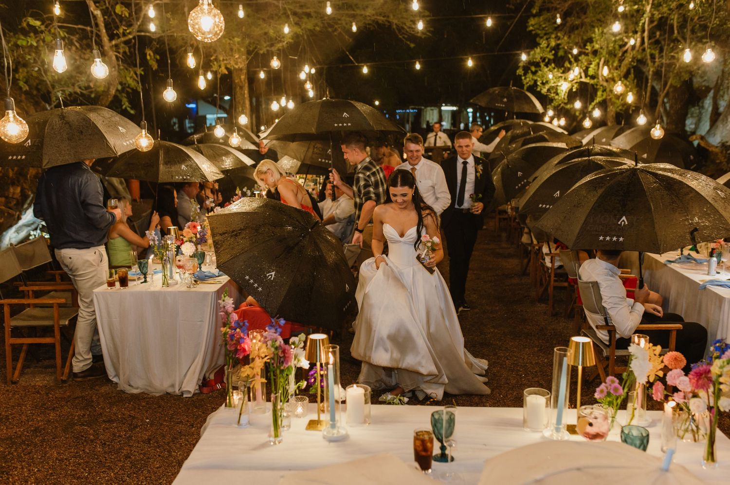 Wedding guests holding umbrellas in rain at outdoor reception with string lights and colorful floral centerpieces.