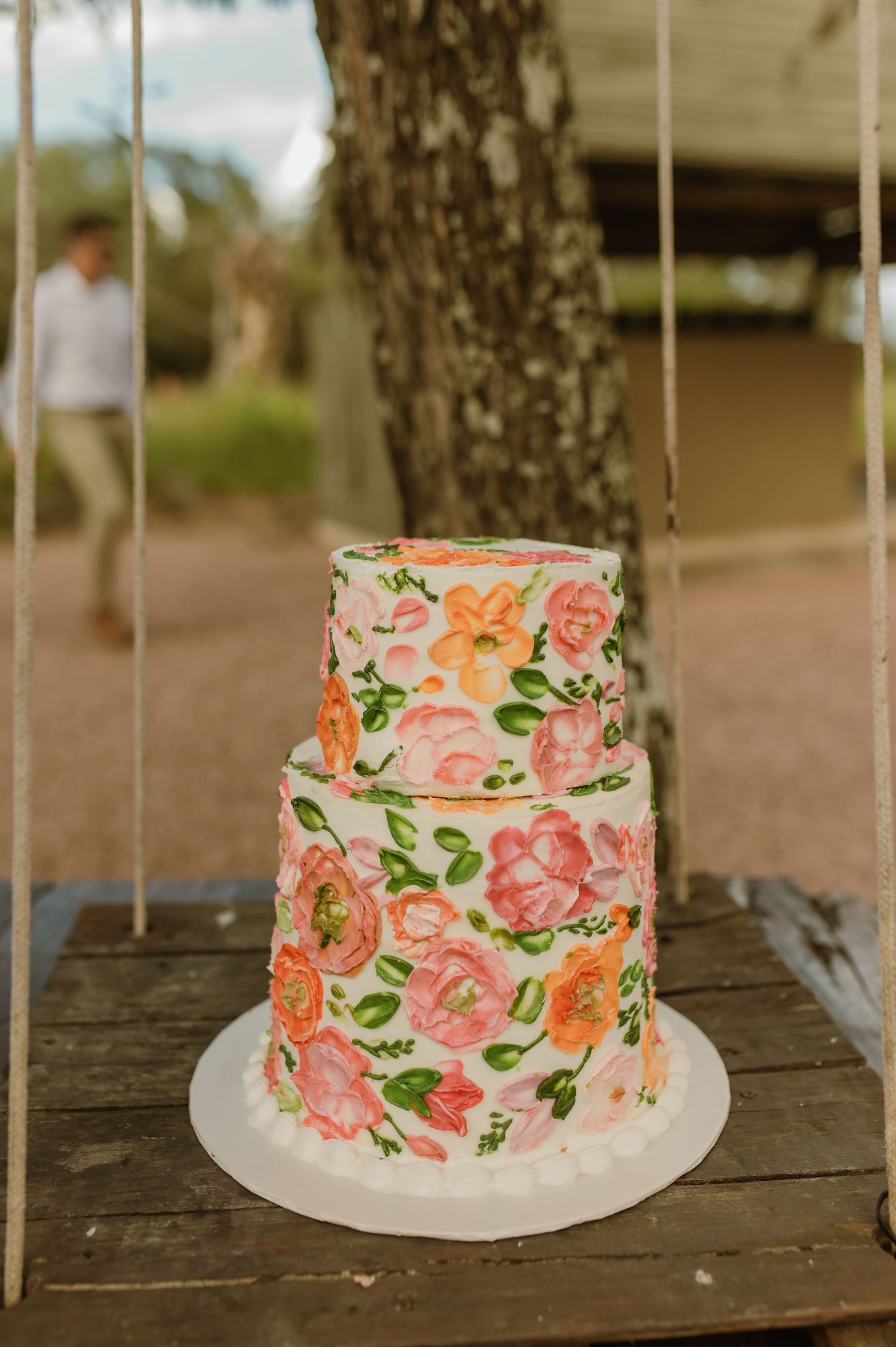 Elegant three-tier white wedding cake decorated with hand-painted pink and orange floral designs outdoors.