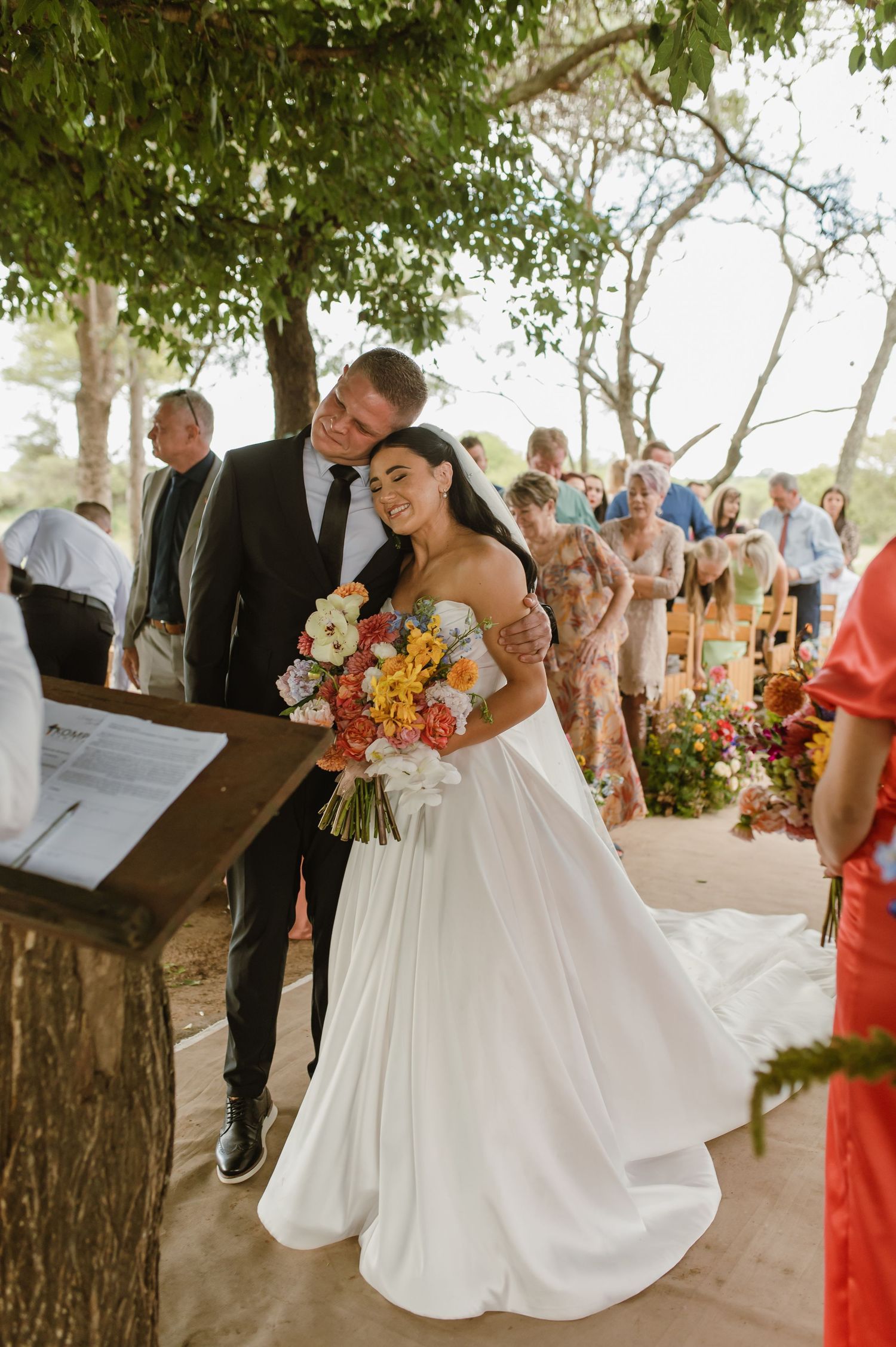 Bride holding colorful bouquet stands with groom at outdoor wedding ceremony altar under lush green trees.