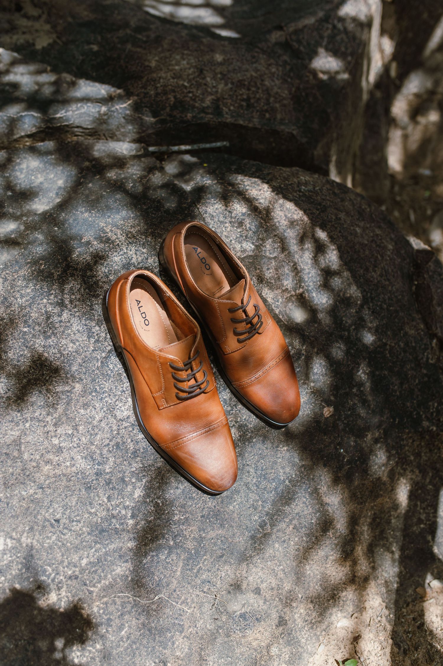 Pair of tan leather dress shoes photographed on stone surface with dappled shadows.