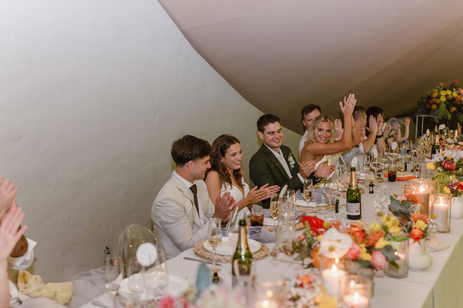 Wedding reception guests enjoy dinner at elegantly decorated tables under string lights in an outdoor tent setting.