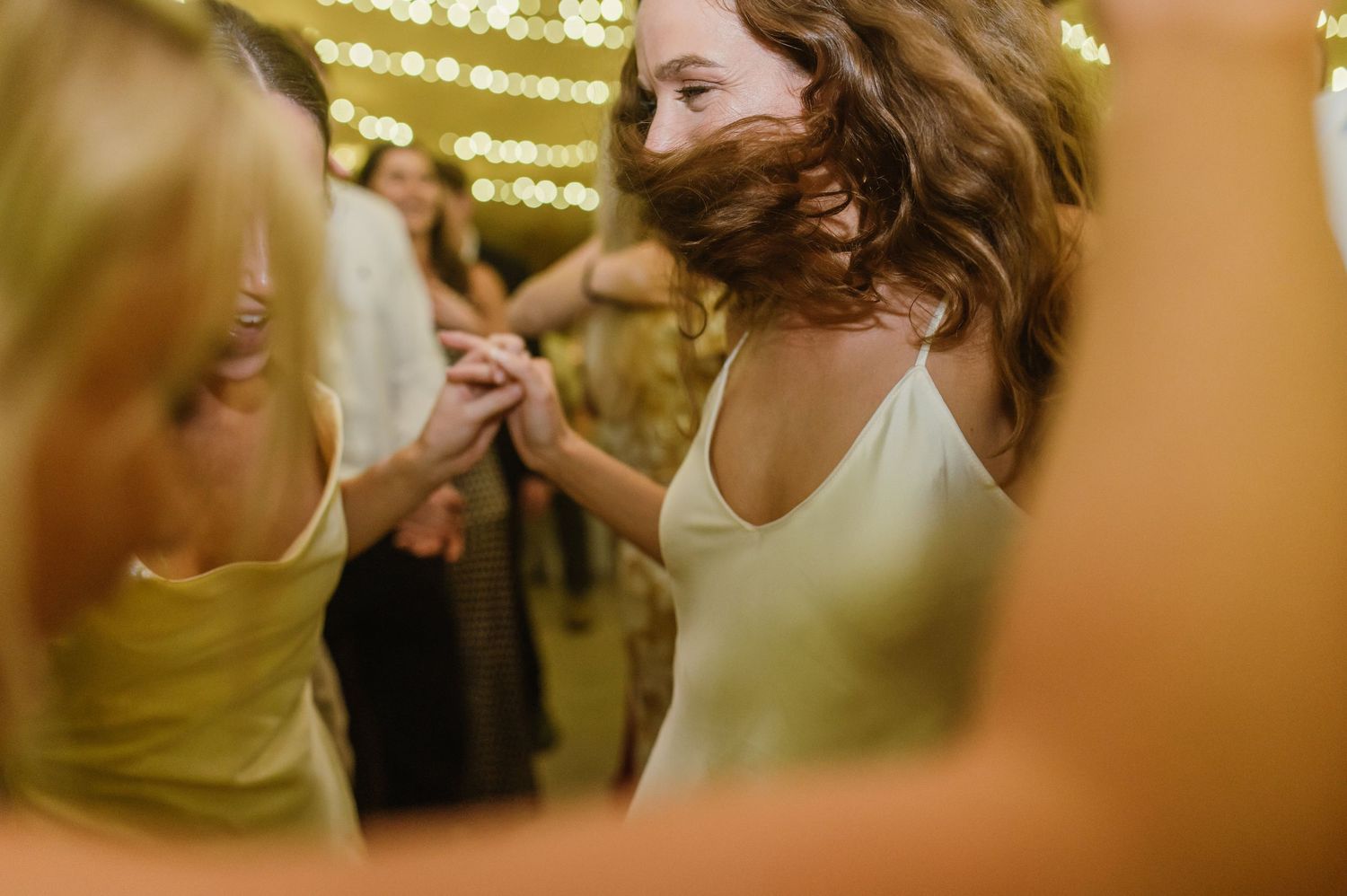 Wedding guests dancing and celebrating beneath glowing string lights at an indoor reception.