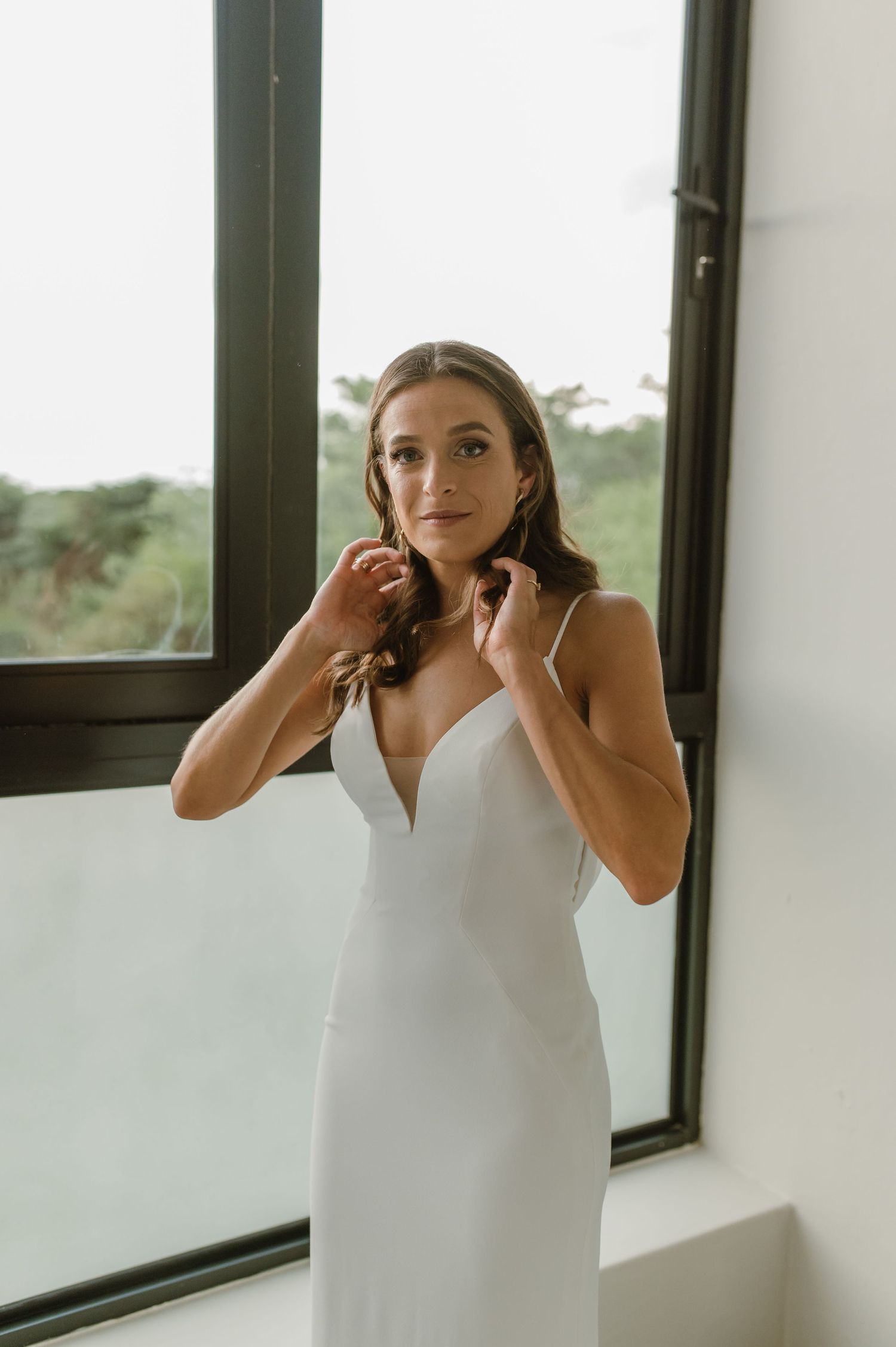 An elegant bridal portrait by a window in a fitted white wedding gown.