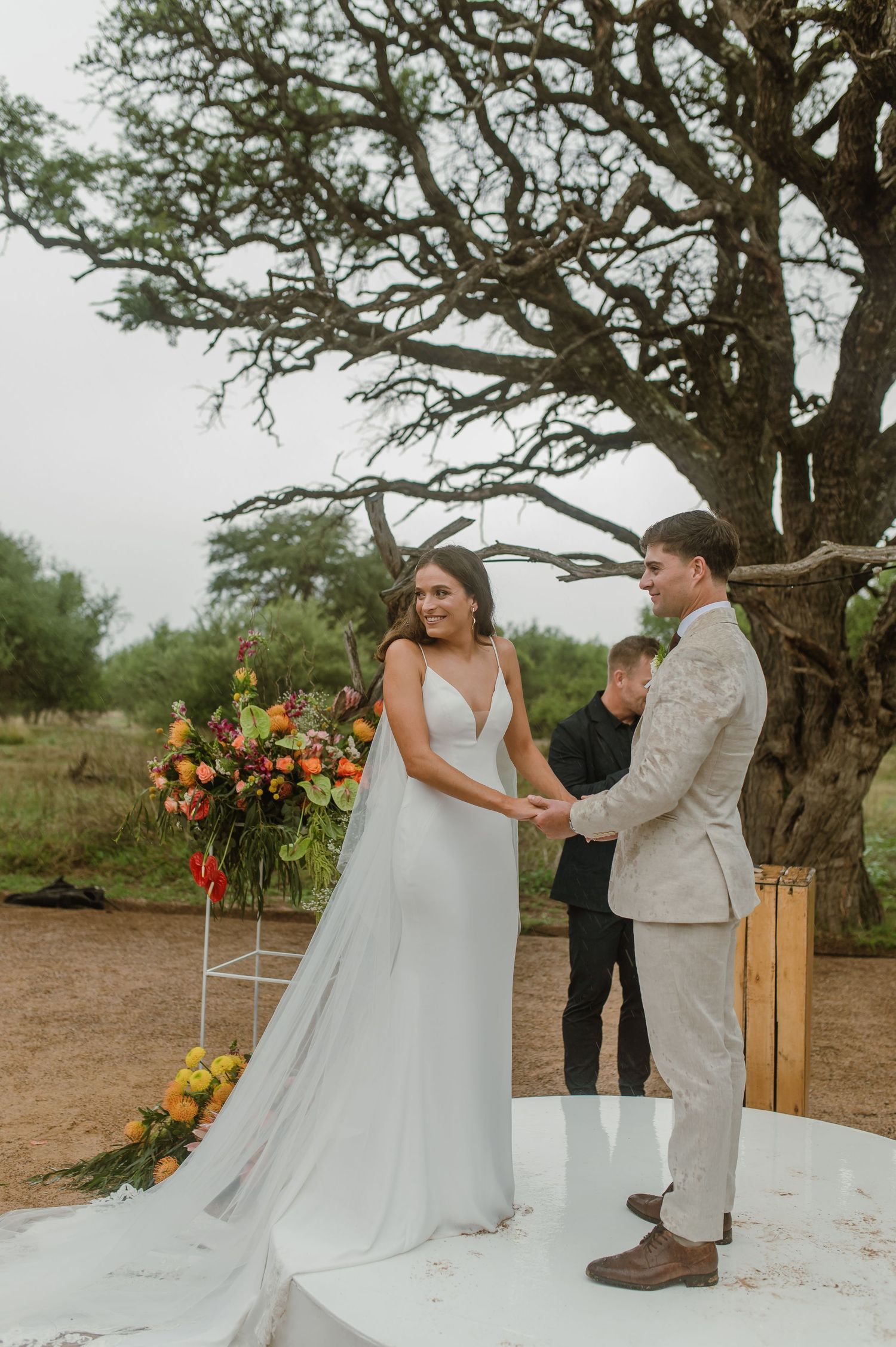 A wedding ceremony takes place on a white platform with vibrant orange and yellow floral arrangements.