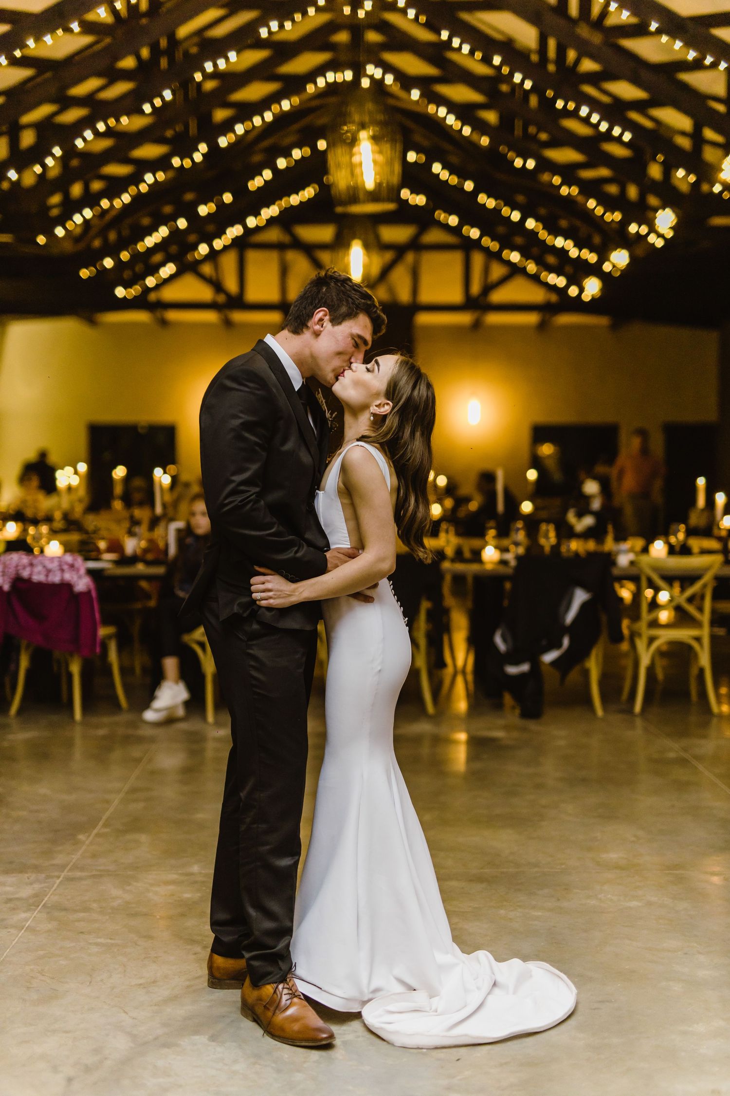A romantic moment between newlyweds under string lights in a rustic venue.