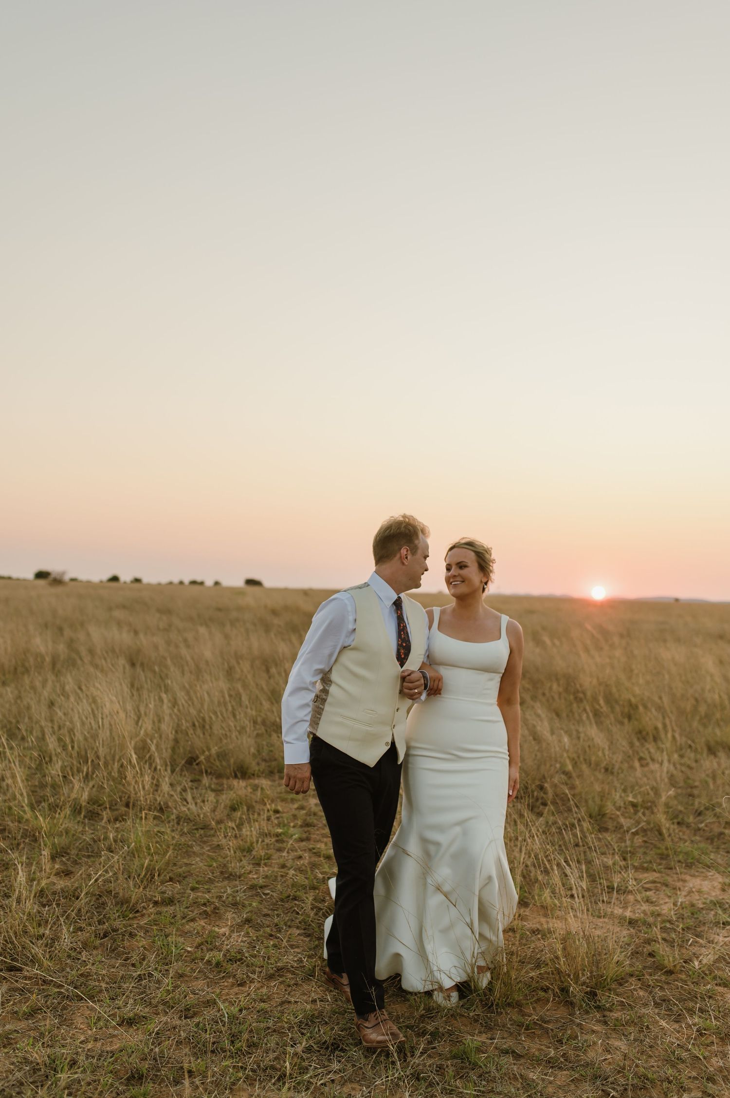 A romantic couple walks through a golden field during sunset.