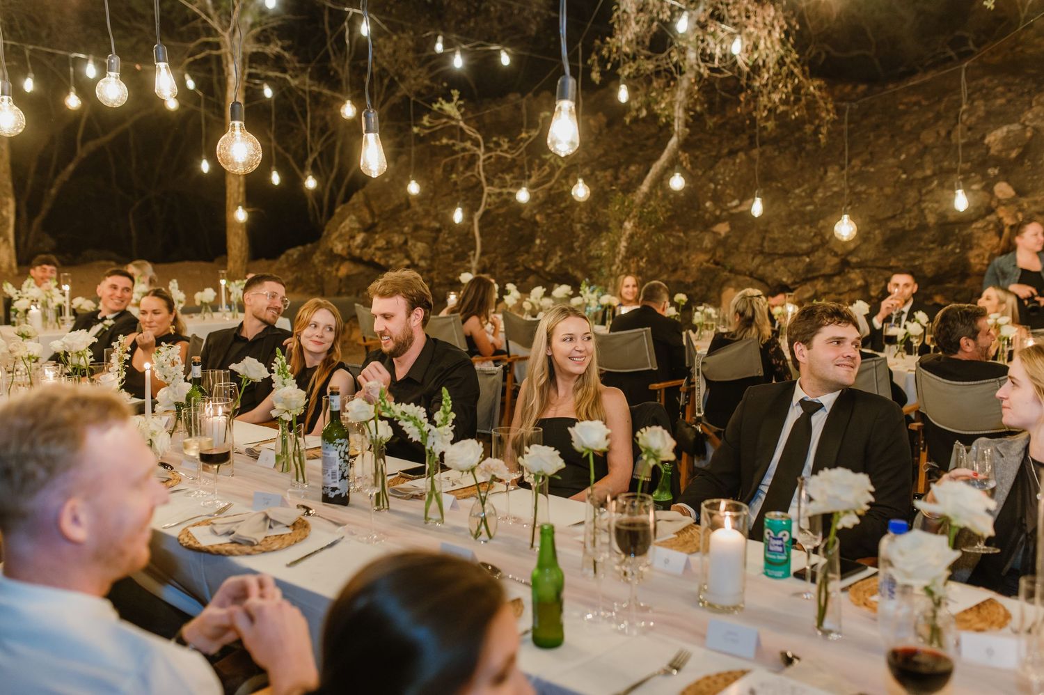 Wedding guests dining at tables under string lights during outdoor evening reception.