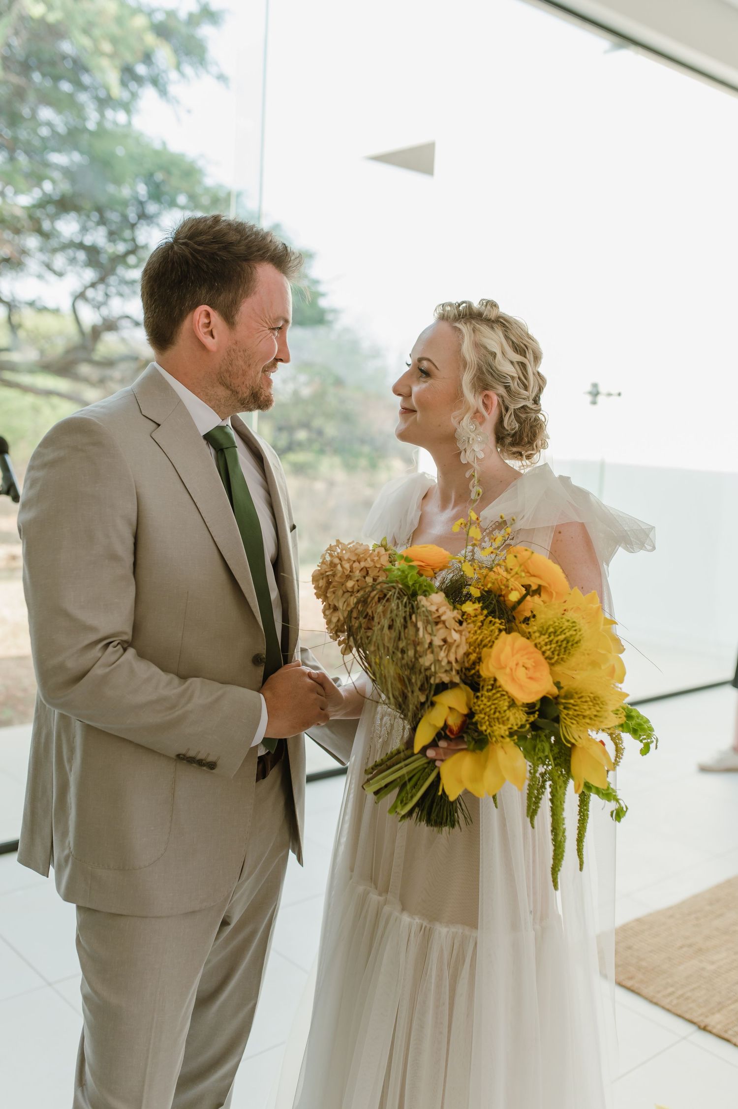 Couple holding bright yellow sunflower bouquets during their first look in a light-filled room.