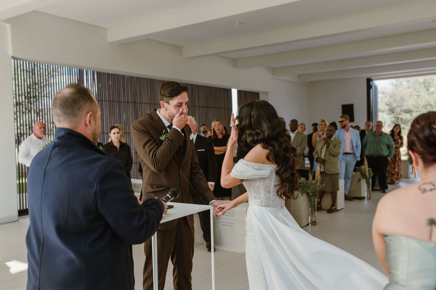 Wedding party members in sage green dresses and dark suits gather for a first look reveal in a modern white venue.