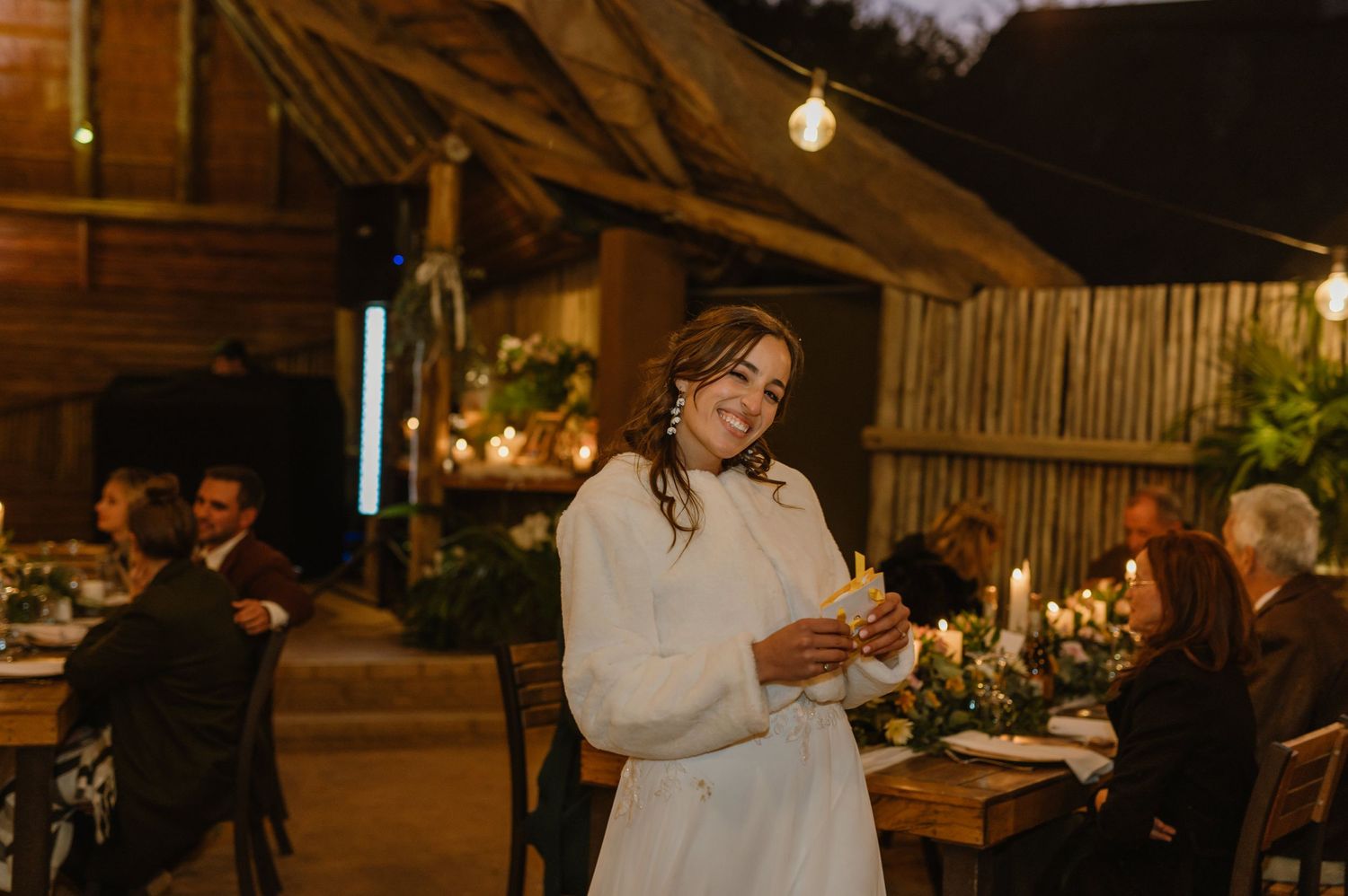 Wedding guests enjoy dinner and conversation at long wooden tables in a warmly lit rustic venue.